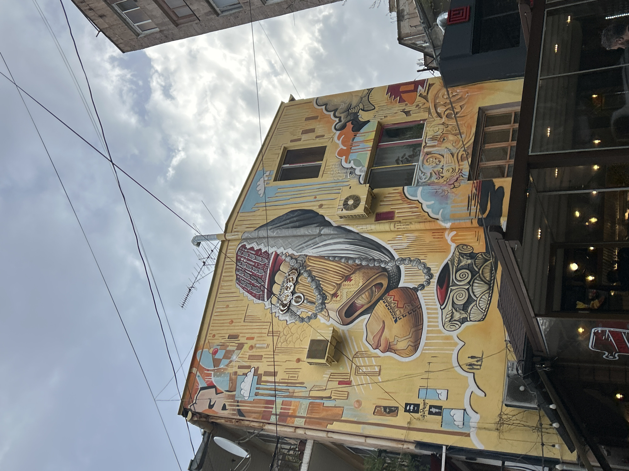The image captures a street scene in Yerevan, Armenia, focusing on a building adorned with a large mural. The mural, painted on the yellow facade of the building, depicts an abstract, stylized figure with intricate patterns and shapes. The mural is a prominent artistic element in the urban environment.
The building has windows and an air conditioning unit, which are part of the overall scene. Overhead, utility wires cut across the sky, adding to the urban atmosphere.
To the right of the mural, a modern structure with large glass windows is visible. This structure appears to be a restaurant or cafe. The image is shot from a low angle, looking upward towards the building. The sky is overcast with cloudy conditions, indicating daylight.