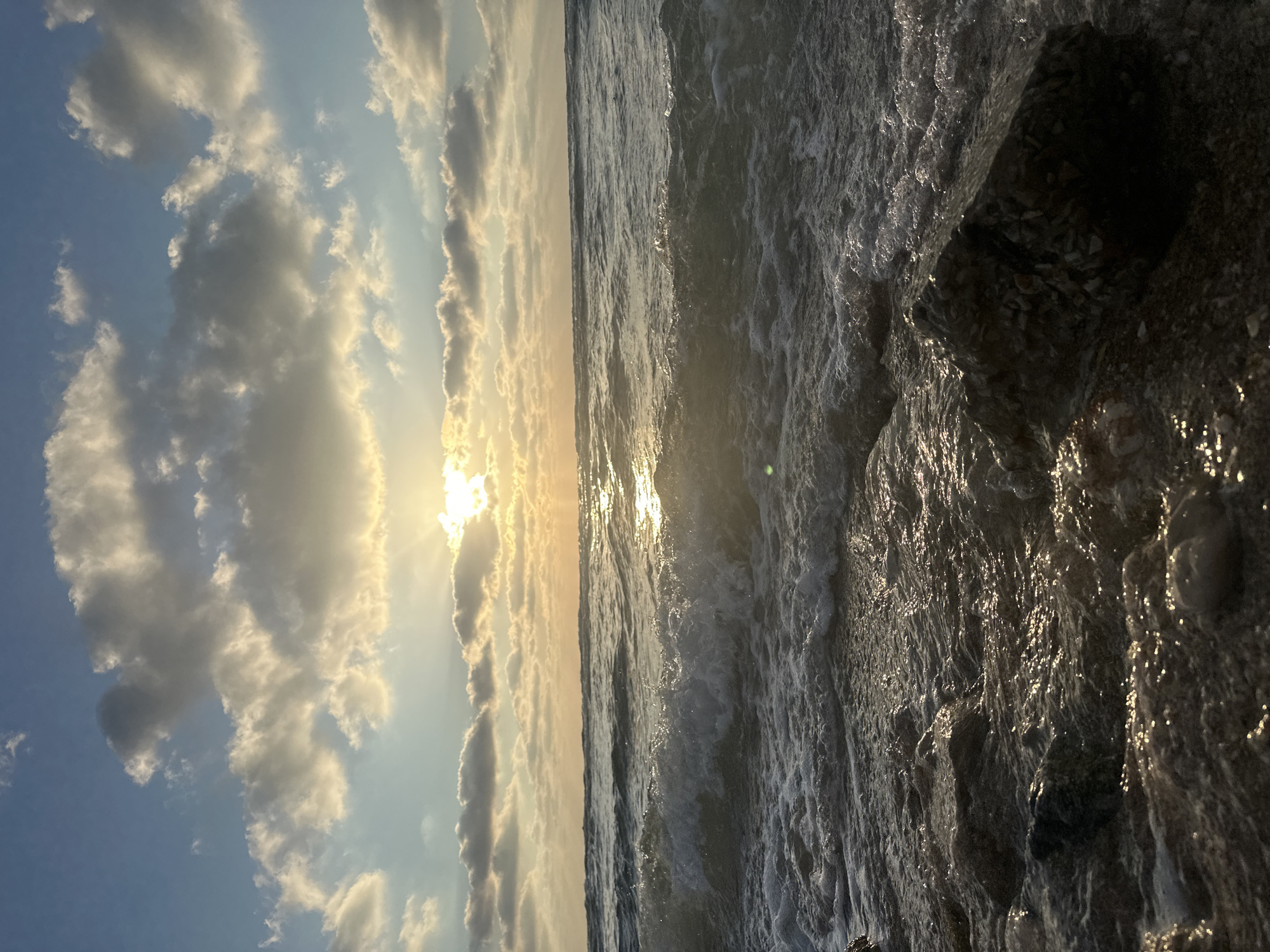 The image captures a sunset over the sea, with the sun partially obscured by clouds, casting golden light onto the water. Waves gently lap against a rocky shoreline in the foreground. The sky is a mix of blue and warm hues, with scattered clouds. The scene is peaceful and serene, evocative of a quiet evening on the coast. The location is the city of Haifa, Israel, specifically its Mediterranean coastline. There are no people visible in the image.