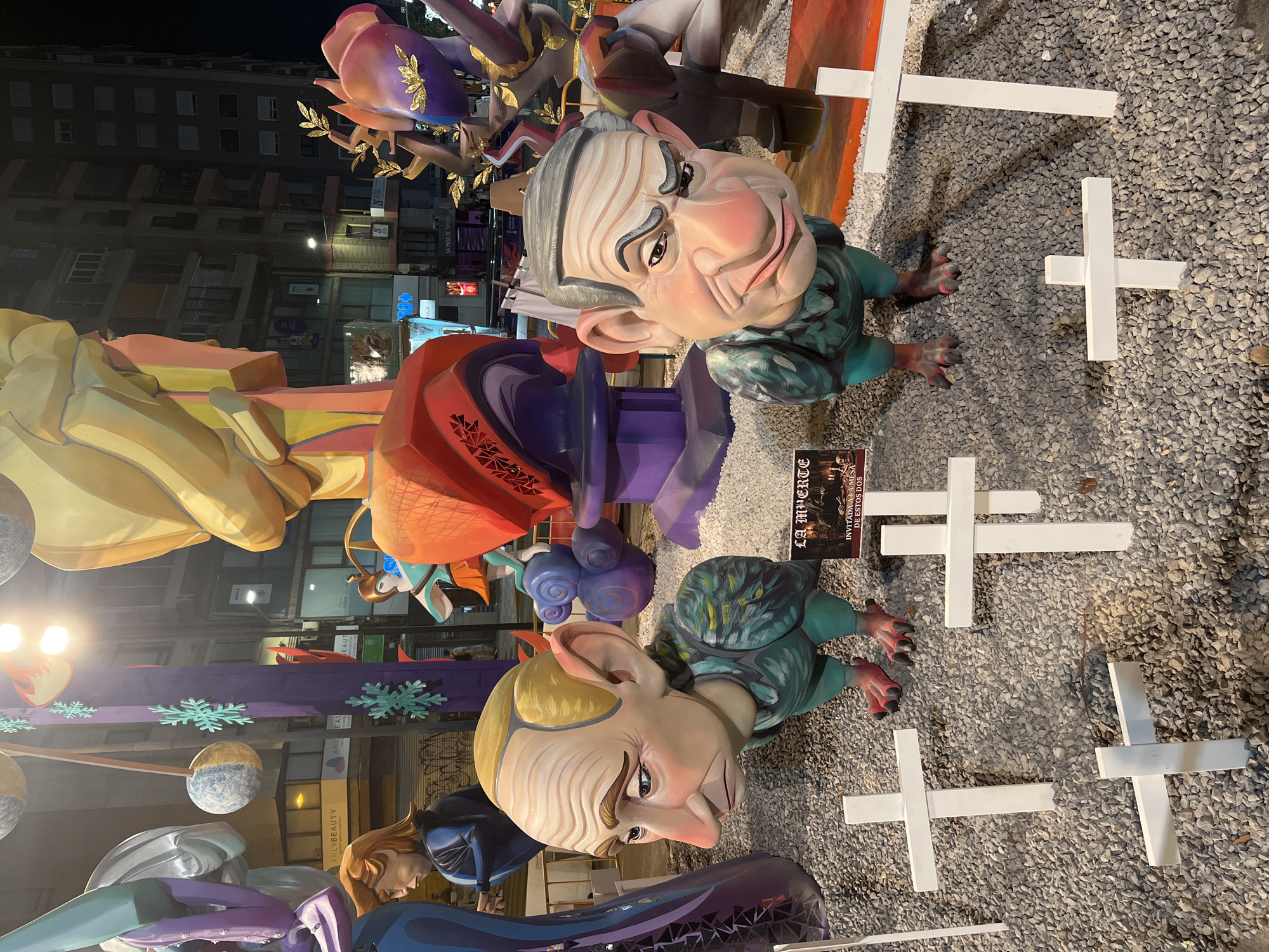 The image depicts a nighttime scene in Alicante, Spain, featuring large, cartoonish effigies and decorative elements. In the foreground, two prominent figures with exaggerated features stand amidst white wooden crosses placed on a gravel ground. One figure, on the left, has a slumped posture and a pensive expression, with pale skin, blond hair, and large ears. It wears a teal, feathery garment. The figure on the right is more alert, with graying hair, a stern expression, and a similar teal garment. Between these figures, a small sign reads "LA MUERTE" with further text below. Behind them, other colorful, abstract sculptures and figures are visible, along with building facades and streetlights in the background. The overall impression is of a festive or artistic display, possibly a "Falla" monument, common in Valencian communities including Alicante.