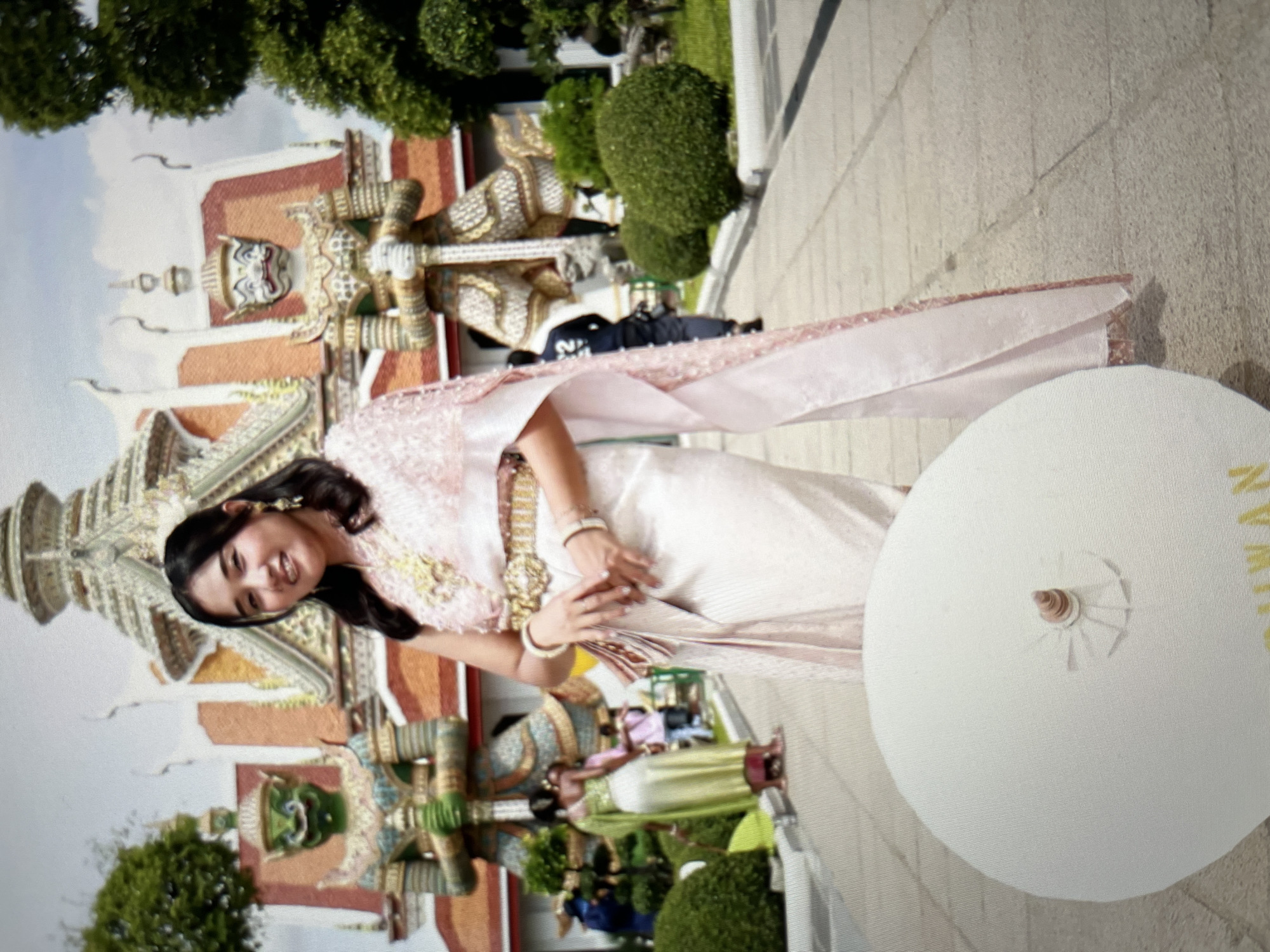 A young woman in traditional Thai attire, featuring a pink and gold embroidered top, a pale pink silk skirt, and a sheer pink shawl, stands in front of a temple with large, ornate statues of guardian figures. She is smiling and holding a white umbrella in her left hand, with her right hand resting on her skirt. The temple has a distinctive tiered roof, and the guardian figures are imposing with green and white faces. In the background, other people are visible, some in traditional dress and others in casual clothing, walking along a paved pathway. Green trees and manicured bushes add to the outdoor setting. The umbrella in the foreground has some yellow text that is partially obscured. The overall scene is a cultural and historical landmark, likely in Thailand. This image is not located in Cirahayu, Indonesia.