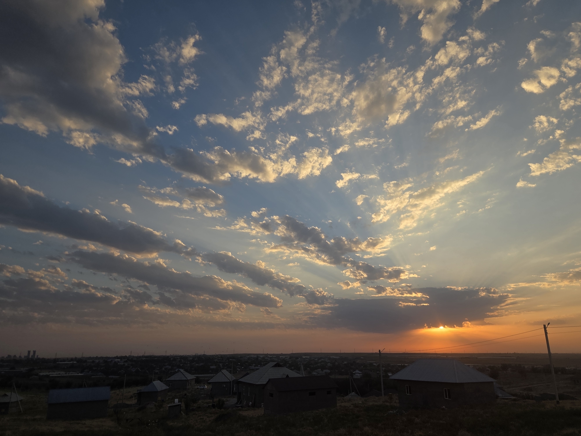 The image depicts a sunset over a residential area. The sky is filled with clouds illuminated by the setting sun, which is visible on the right side of the horizon, casting orange and yellow hues. Rays of light break through the clouds, creating a dramatic effect. Below the sky is a view of a town or village, with numerous small houses, some with distinctive gray roofs. The terrain in the foreground is uneven and covered with dry grass. In the distance, a hazy cityscape or industrial area can be seen. The scene is set in the city of Qarasy, Kazakhstan. There are no visible people or animals in the image. The overall impression is one of a rural or suburban landscape at dusk.