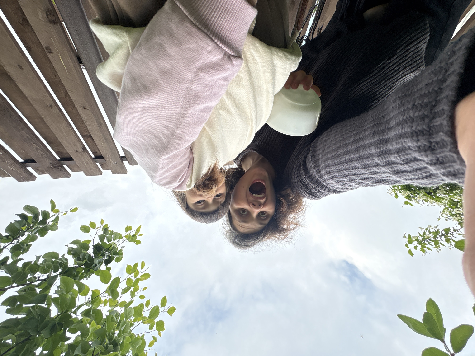 Two individuals are visible in the image. One appears to be a younger child with brown hair, and the other is an adult with brown hair, who is opening her mouth widely. The individuals are positioned under a wooden structure, possibly a deck or overhead walkway. The background includes foliage, with green leaves and branches visible. The sky is also visible between the branches, indicating an outdoor setting. One of the individuals is holding a white object. It is impossible to confirm the location in Прикамский, Russia.