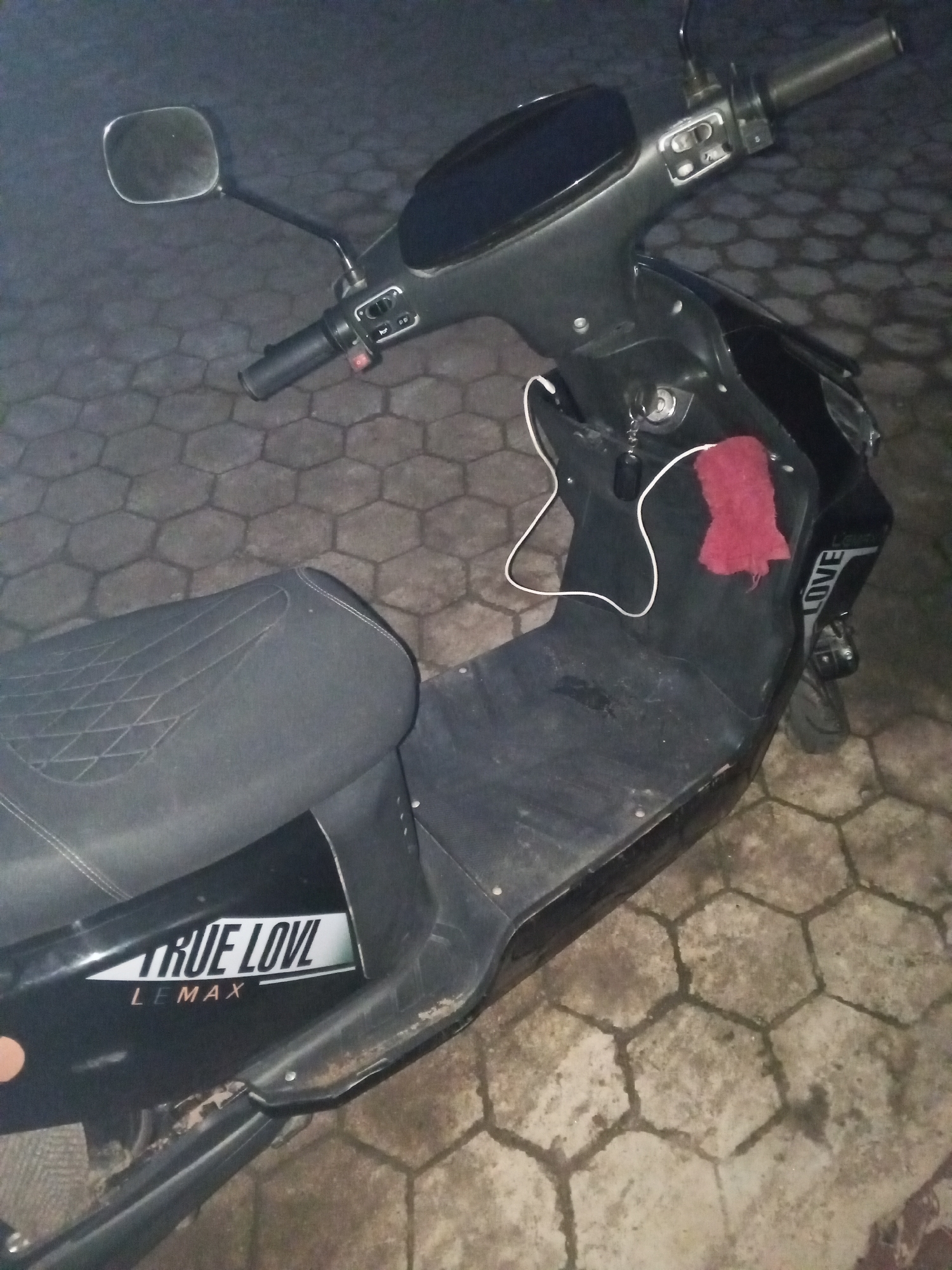 An electric scooter is parked on a hexagonal tiled surface in an outdoor environment, likely a street or parking lot in Jos, Nigeria. The scooter is black with a textured, diamond-stitched seat and a prominent "TRUE LOVE LEMAX" decal on its side. A red cloth is draped over the scooter's body. The scooter has a visible mirror on the left side and appears to be in a parked state. The lighting suggests it is nighttime or very early morning, with the tiled ground showing some dampness or discoloration. There are no people visible in the image, and no apparent actions or interactions are taking place.