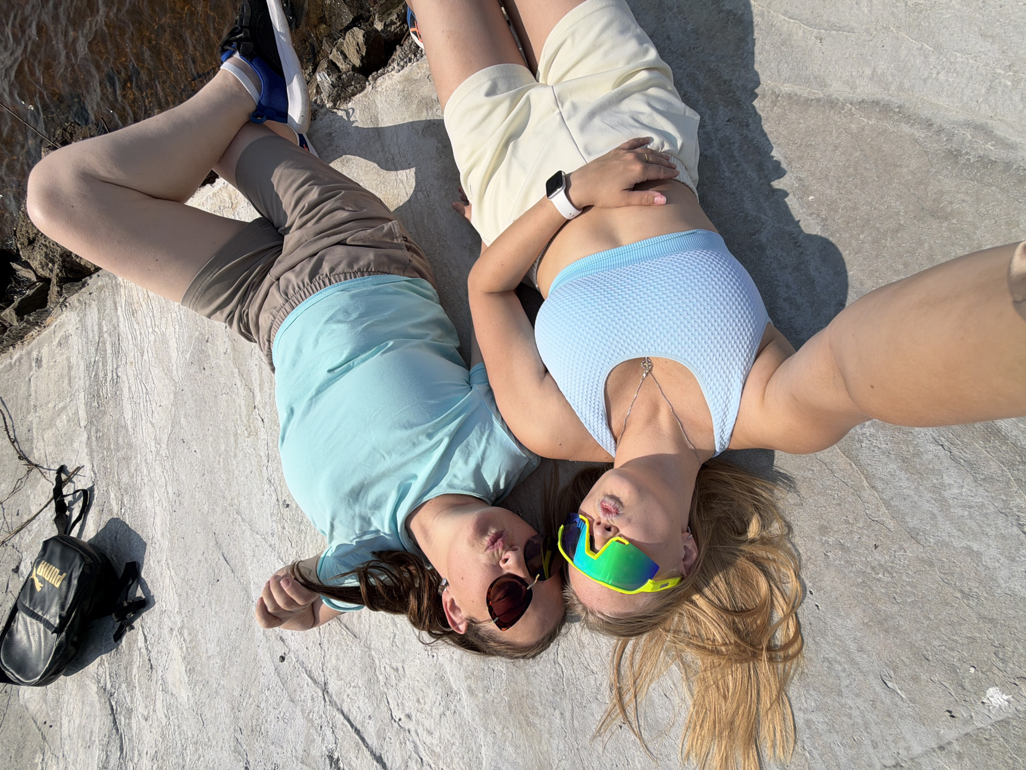 Two individuals are lying on a textured concrete surface. The person on top is wearing a light blue ribbed sports bra and light beige shorts. They have long blonde hair and are wearing neon green and blue wrap-around sunglasses. The person below is wearing a light turquoise t-shirt and taupe shorts. They have brown hair tied in a braid and are wearing dark brown, oversized sunglasses. Both individuals are making duck faces, suggesting a posed photograph. A black backpack with gold lettering is visible on the ground to the right of the individuals. The environment appears to be outdoors, with shadows cast by the subjects, indicating sunlight. The concrete surface appears to be part of a pathway or embankment. The presence of water and rocks near the edge of the concrete suggests the location might be a waterfront area. Based on the image alone, it is impossible to definitively determine the specific location within Chaikovsky, Russia. However, the visual elements suggest a recreational area near a body of water.