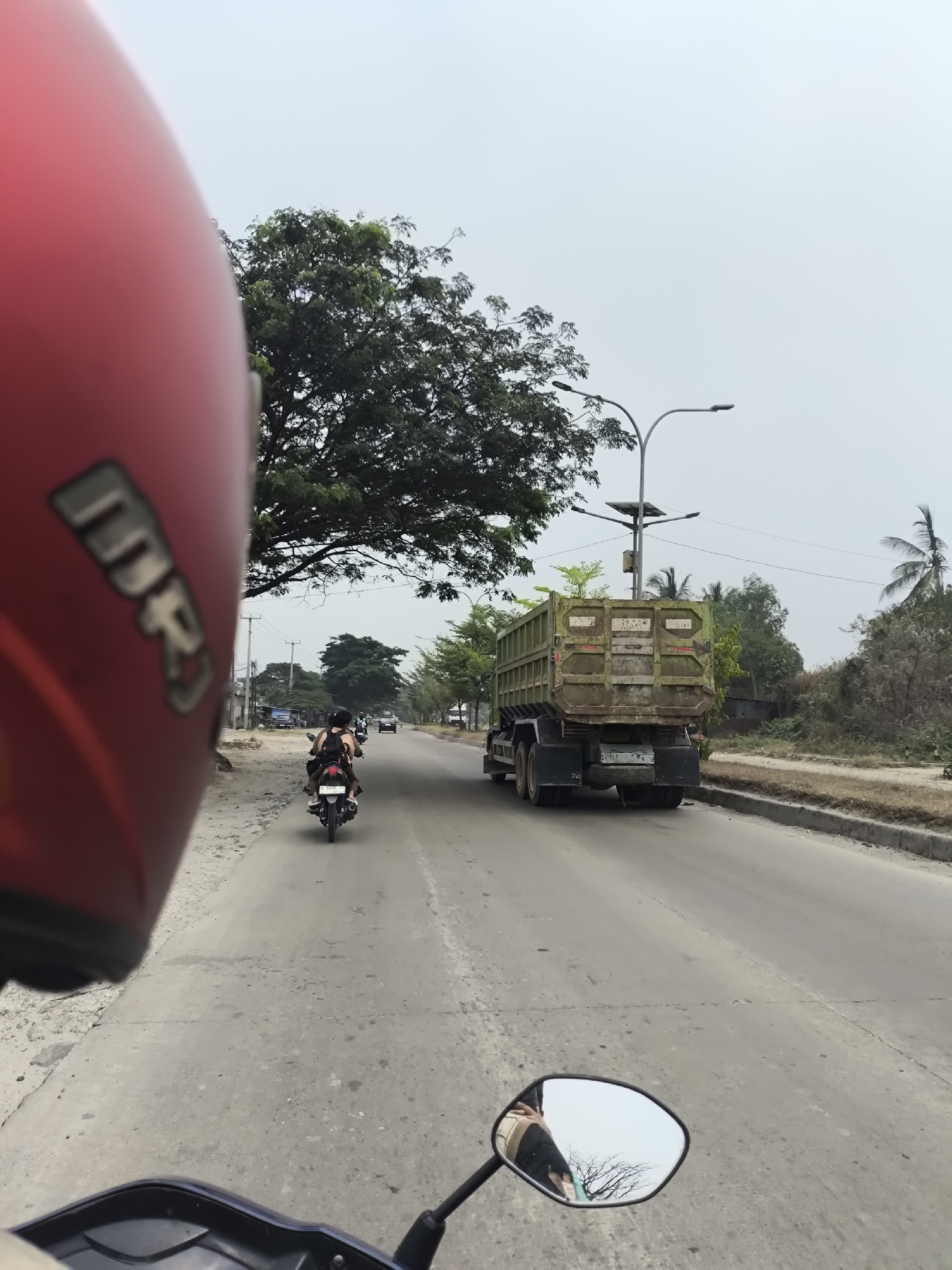 A first-person perspective captures a view from a motorcycle on a two-lane road in Cilegon, Indonesia. The image is dominated by a red motorcycle helmet in the foreground, obscuring the left side of the frame. In the distance, a green dump truck is traveling in the same direction as the motorcycle. Two people are riding on a scooter ahead of the dump truck, and other vehicles and trees line the roadside. The sky is overcast, suggesting a cloudy day.