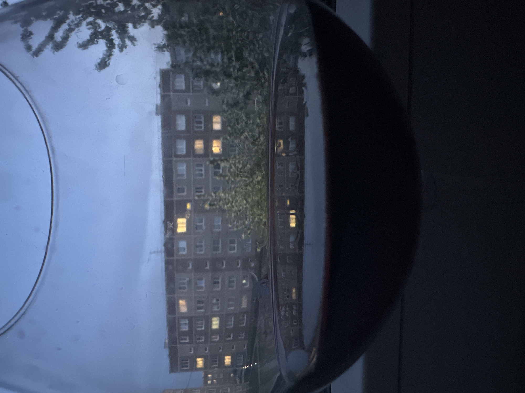 The image shows a view through a glass bowl, distorting a cityscape outside. The focus is primarily on a large residential building in the distance, its windows illuminated with a yellow-orange glow, suggesting it's evening or dusk. A tree partially obscures the building. The bowl itself is slightly out of focus, with water droplets visible on its surface. The right side of the image is dark, showing part of a window frame or wall, creating a contrast with the brighter cityscape. Given the prompt, the location is presumed to be Chaikovsky, Russia, although no specific landmarks are visible in the image to confirm this.