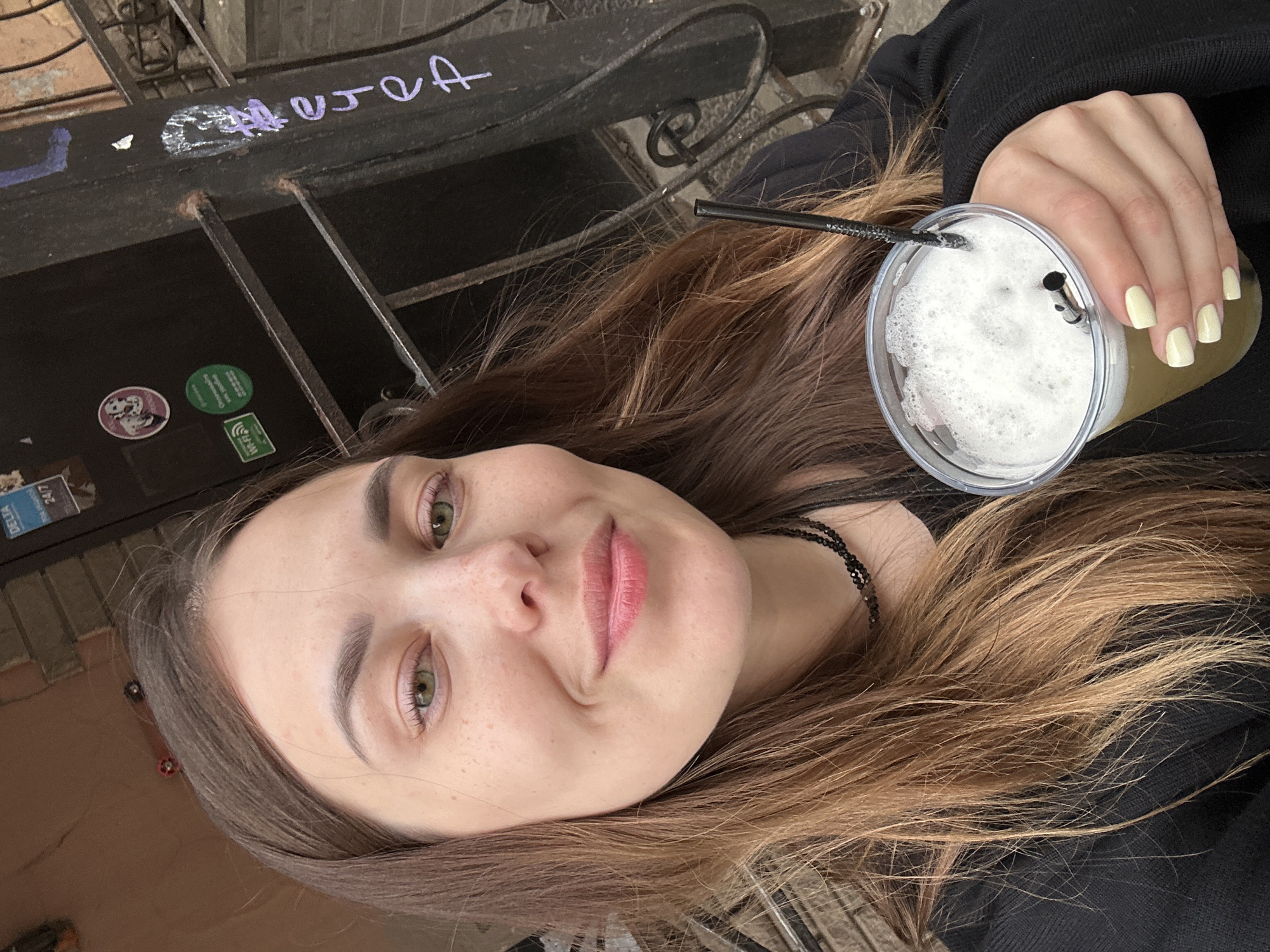 A young woman with long, brown hair and light green eyes is holding a plastic cup of a frothy, light-colored beverage. She is wearing a black top and a black beaded necklace. The background appears to be an urban setting with a brick wall and a metal fence. Graffiti is visible on a dark metal pole. There are stickers on a dark door in the background, including a Wi-Fi symbol and what appears to be a "Delta 24/7" sticker. The overall impression is of a casual, outdoor snapshot.