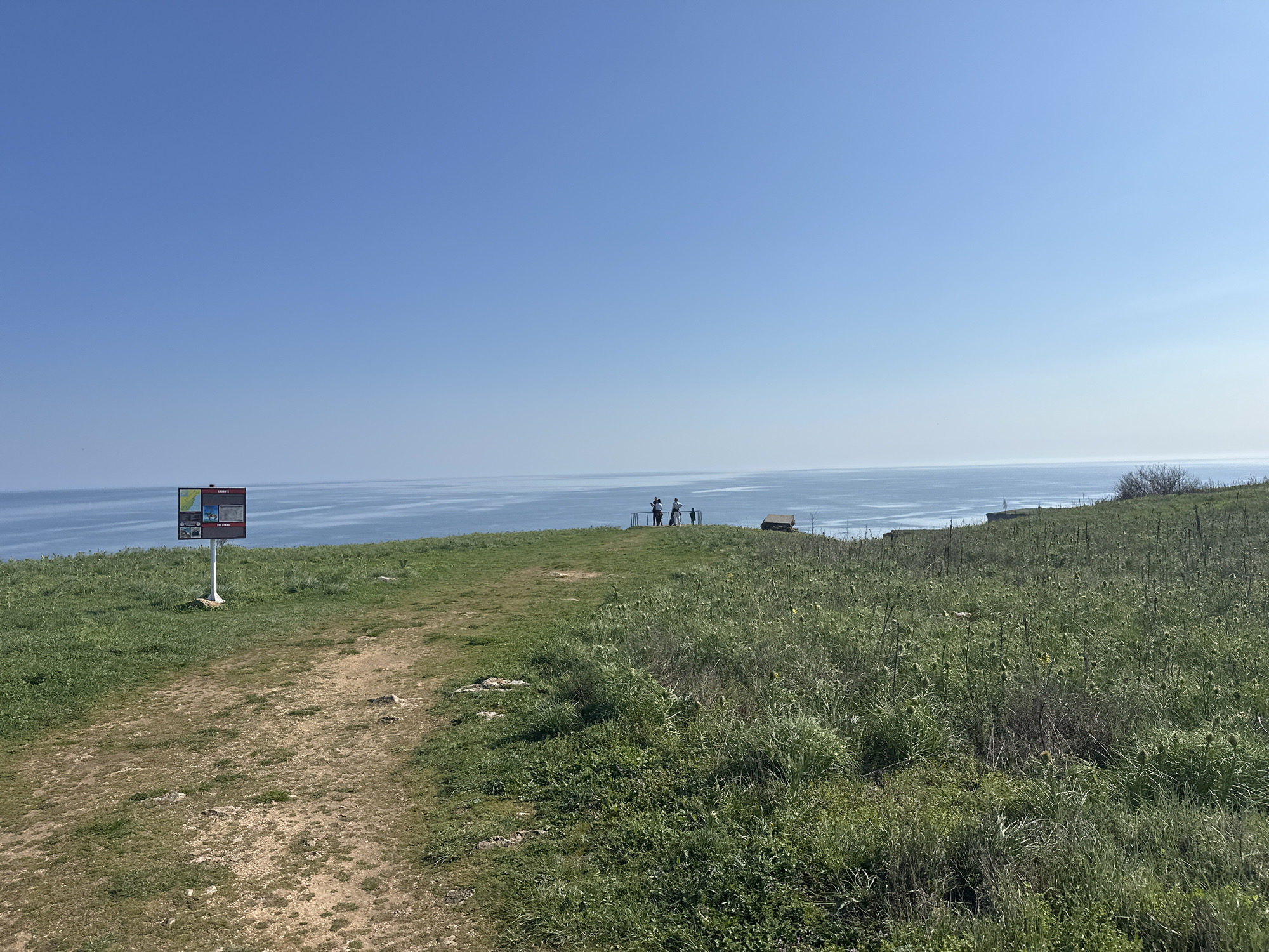 The image shows a coastal landscape in Kamen Bryag, Bulgaria. A dirt path leads from the foreground towards the edge of a grassy cliff overlooking the Black Sea. To the left of the path is an informational sign on a post. At the cliff's edge, a group of three or four people stand looking out at the sea. A small, low structure is visible near the group. The sky is clear and blue. Vegetation covers the ground.