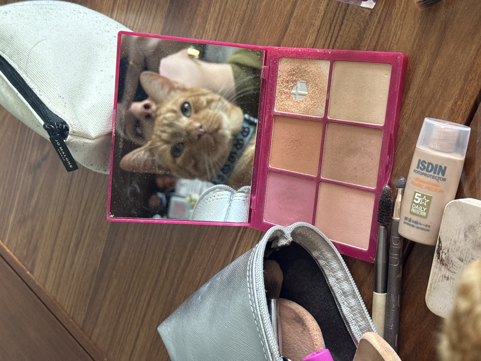 On a wooden surface, likely a table, several cosmetic items are arranged. These include a white zip-up pouch with "JO MALONE" printed on the zipper tag, a pink makeup palette with six different shades, a tube of "ISDIN Fotoprotector Fusion Water Magic Medium", and a silver cosmetic bag containing makeup brushes. The mirror within the makeup palette reflects an orange cat wearing a blue collar, along with a person's face and hand holding the palette. The scene is likely indoors. It is not possible to discern the location of the city of Mexico City, Mexico.