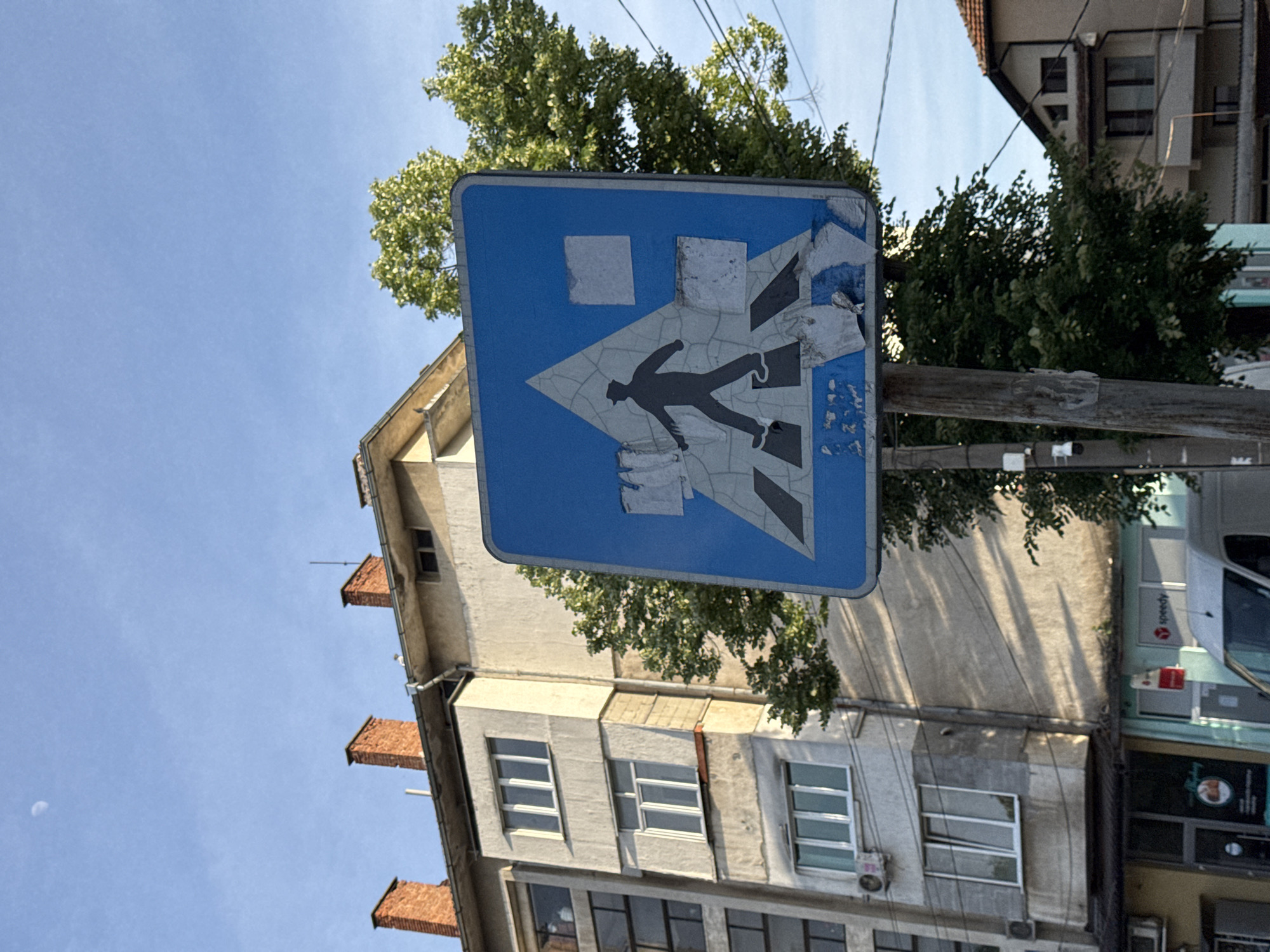 A pedestrian crossing sign in Lovech, Bulgaria, is affixed to a weathered wooden pole. The blue sign has white markings indicating a pedestrian crossing, though portions of the markings are peeling. The sign is set against a backdrop of a multi-story building with a mix of old and newer architecture, featuring brick chimneys, windows, and light-colored walls. Green trees are visible behind the sign and the building. The sky is light blue. Part of a delivery vehicle can be seen to the right. Power lines crisscross above.