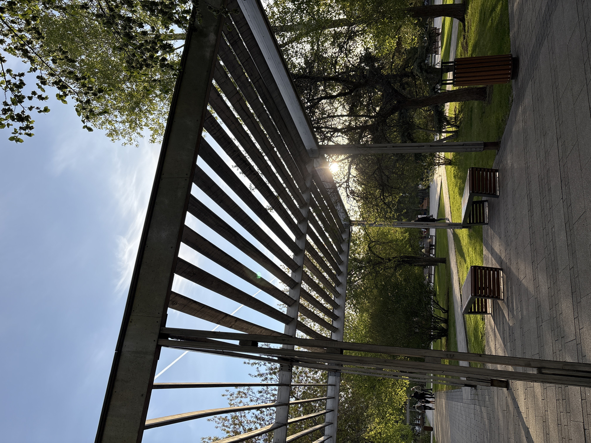 A low-angle, outdoor photograph shows a pergola constructed of evenly spaced, parallel, horizontal slats supported by metal posts. The pergola is oriented diagonally across the frame, positioned above a paved walkway with grass and trees on the right side. Sunlight streams through the pergola slats and tree branches.
On the walkway, three benches and two waste bins are visible. Beyond the walkway, a grassy area stretches back to a row of trees in the background. The sky is blue with thin clouds and contrails visible through the slats of the pergola. Several people are walking under the pergola.
The photograph was taken in Krasnoyarsk, Russia.