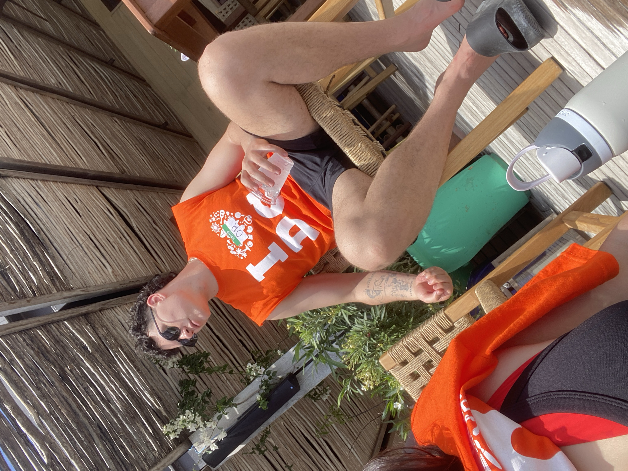 The image shows a man wearing an orange "HELLO OTTAWA" shirt, black shorts, and sunglasses. He is sitting on a woven chair, holding a glass of water. A water bottle and a green cushion are next to him. He has a tattoo on his left arm. In the background, there is a thatched roof, suggesting a beach or resort setting. White flowers and green plants are also visible. The location is Marina Maroma, Mexico.