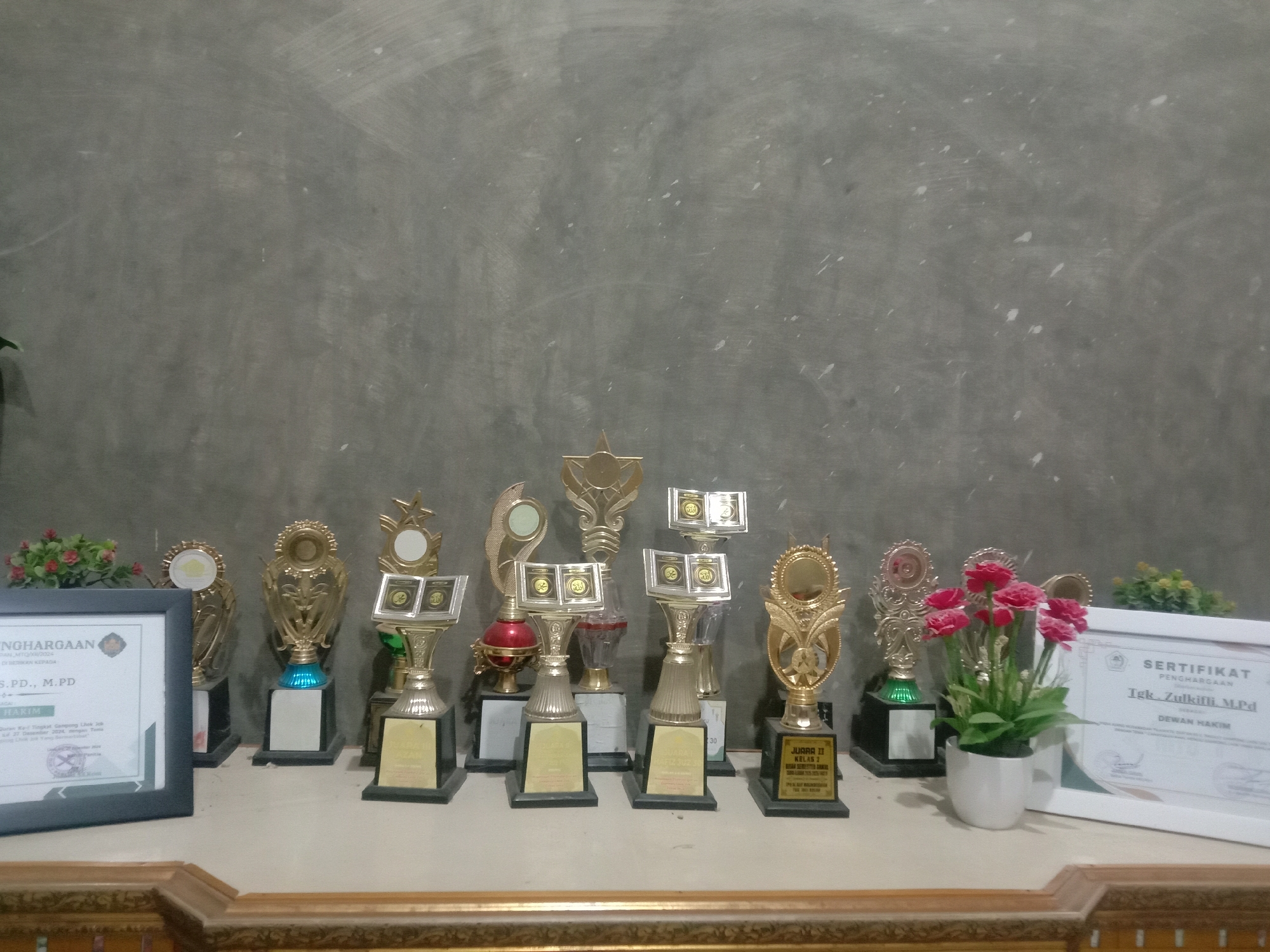 The image shows a collection of trophies and certificates displayed on a shelf. The trophies vary in design, with some featuring open book motifs and others with more traditional shapes. The certificates are framed, one bearing the title "SERTIFIKAT PENGHARGAAN" (Certificate of Appreciation) and the name "Tgk. Zulkifli, M.Pd" with "DEWAN HAKIM" (Board of Judges). Another is visible on the left side of the image. The presence of certificates and the prominence of the "JUARA II" (Second Place) label suggest the trophies are awards. The scene is set against a neutral gray wall. Two pots containing artificial red flowers and green plants are interspersed among the trophies and certificates. The location might be the city of Lhokjok, Indonesia.