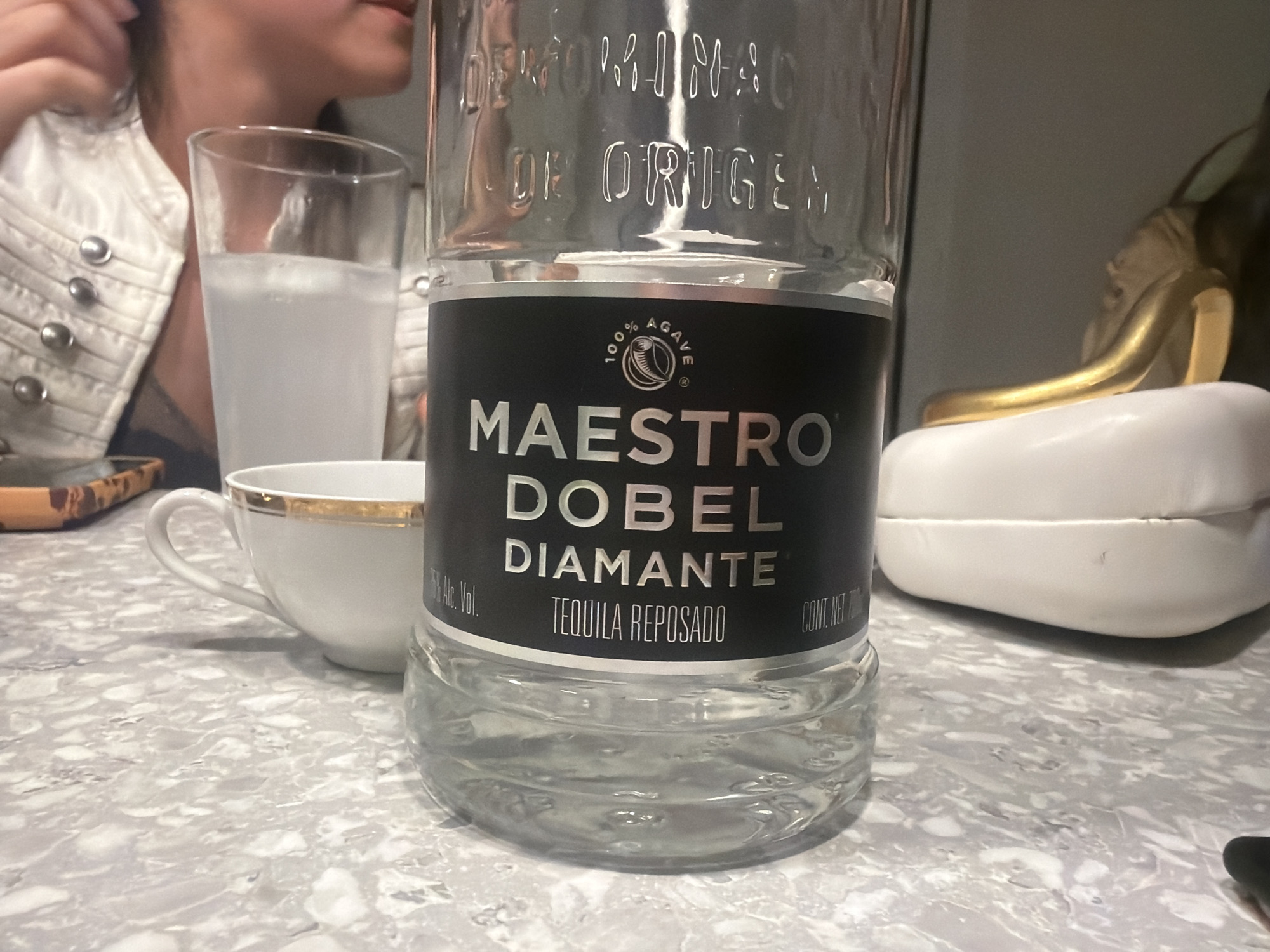 The image shows a partially consumed bottle of Maestro Dobel Diamante Tequila Reposado. The bottle is in the foreground, with its black label prominently displayed. The table beneath the bottle has a light gray and white speckled pattern. Behind the tequila, there is a glass of a light-colored beverage, partially obscured by the bottle. A white teacup with a gold rim is adjacent to the glass. To the right of the bottle is a white object, possibly a clutch, with a gold detail. A person wearing a white jacket with silver buttons is visible on the left side of the frame. The background is indistinct. The location is not discernible from the image, and there is no indication that it is in Milan, Italy.