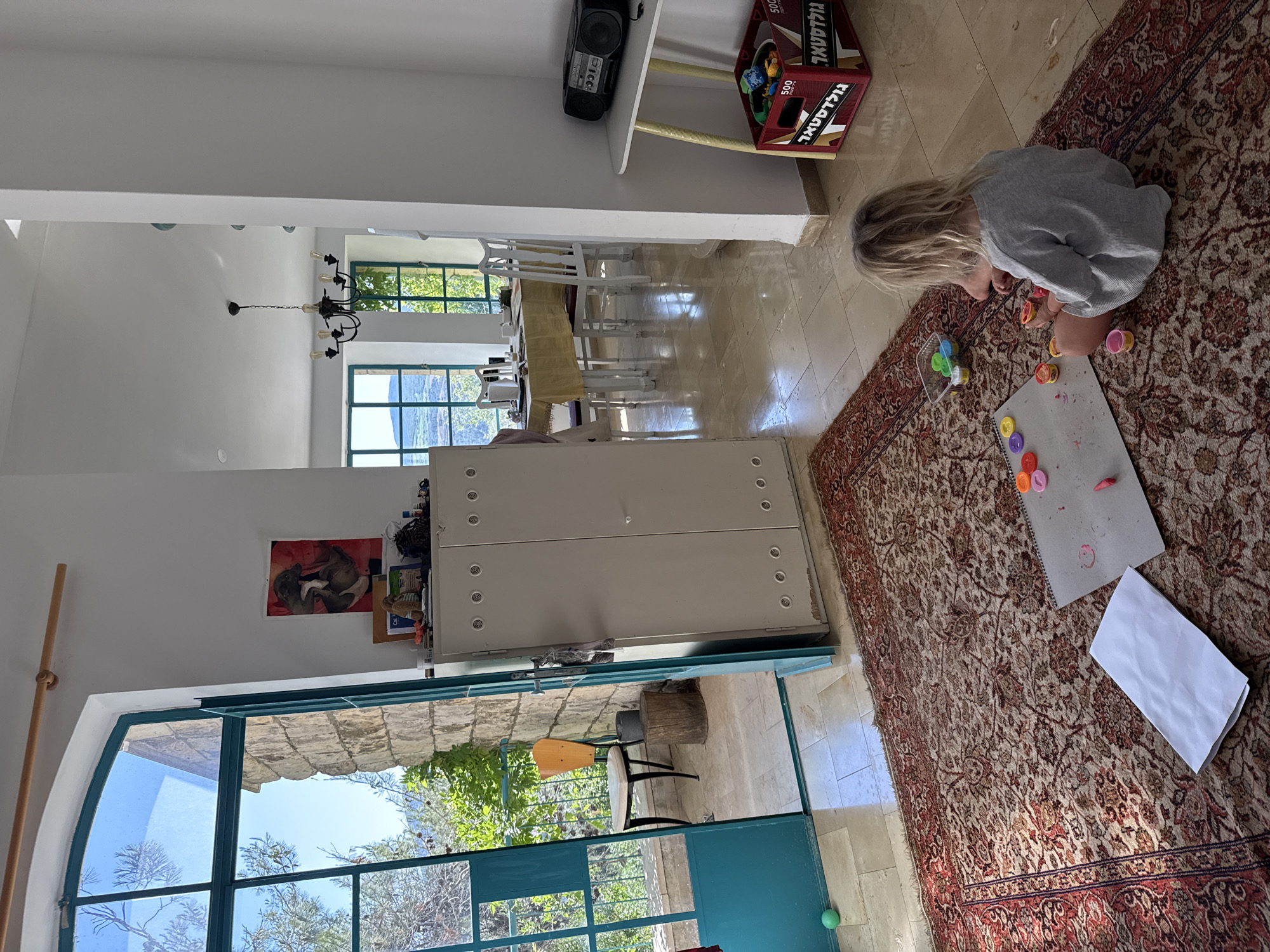 A bright, sunlit indoor scene captures a child engrossed in creative play within a home environment. A young child with light hair, wearing a grey long-sleeved top, is seated on a large, ornate red and beige patterned rug. They are focused on an activity involving colorful small containers, possibly paint or play-doh, arranged on a grey drawing pad that shows smudges of color. A folded white paper rests nearby on the rug.
The room features light-colored tiled flooring and white walls. Large windows with distinctive green frames allow ample natural light into the space and offer views of lush outdoor greenery, trees, and stone architecture, suggesting a sunny day. One window on the left has an arched top.
In the midground, a large white metal cabinet with circular details stands against a wall. Further into the background, a dining area is visible with a table, light tablecloth, and several white chairs, illuminated by a black chandelier. Above a section of the wall, a black boombox with an "ICC" logo rests on a white shelf, next to a red cardboard box. This box has Hebrew text, "גולדטאר" (Goldstar), and "500" printed on it, along with "GOLDASTAR" and "MADE IN CHINA". A picture depicting two figures, possibly animals, hangs on a wall near the cabinet. A small green ball is on the tiled floor near a doorway leading outdoors, where a wooden stool or chair is visible. The bright natural light indicates it is daytime.