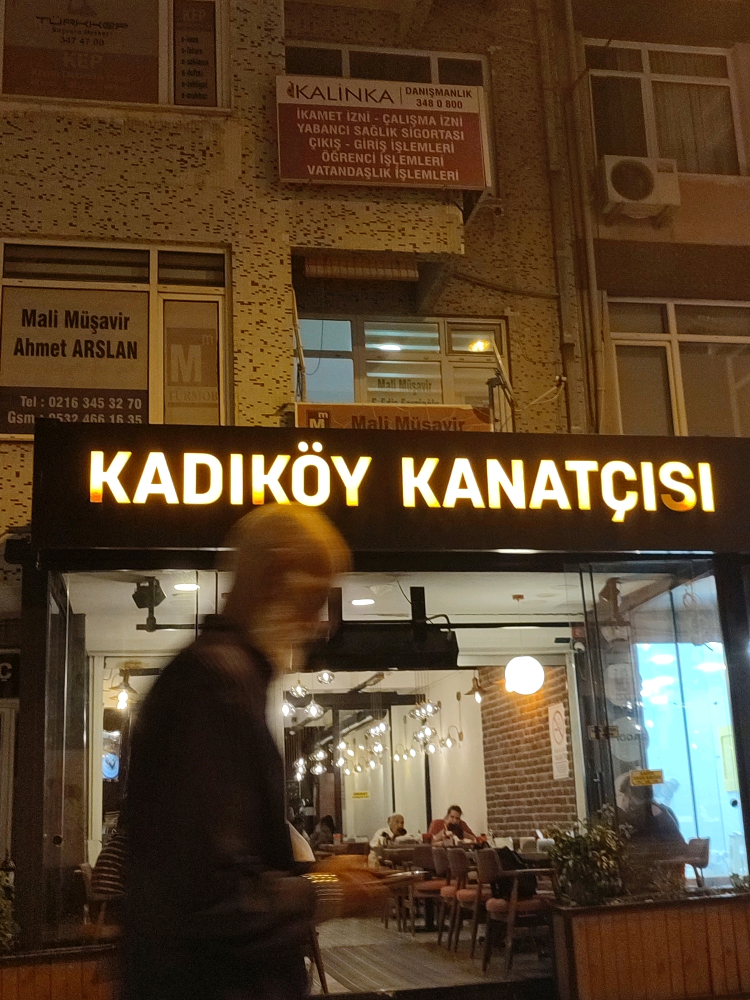 The image shows the exterior of a cafe in Kadıköy, Turkey, at night. A large illuminated sign with the words "KADIKÖY KANATÇISI" is prominently displayed across the storefront. Below the sign, the cafe's interior is visible through a large glass window, revealing tables, chairs, and patrons. A person is captured in motion in the foreground, obscuring part of the storefront. In the background, signs for "Mali Müşavir Ahmet ARSLAN" and "KALINKA DANIŞMANLIK" are visible on the building facade, suggesting the presence of other businesses in the vicinity. The overall scene depicts a bustling urban environment with commercial activity.