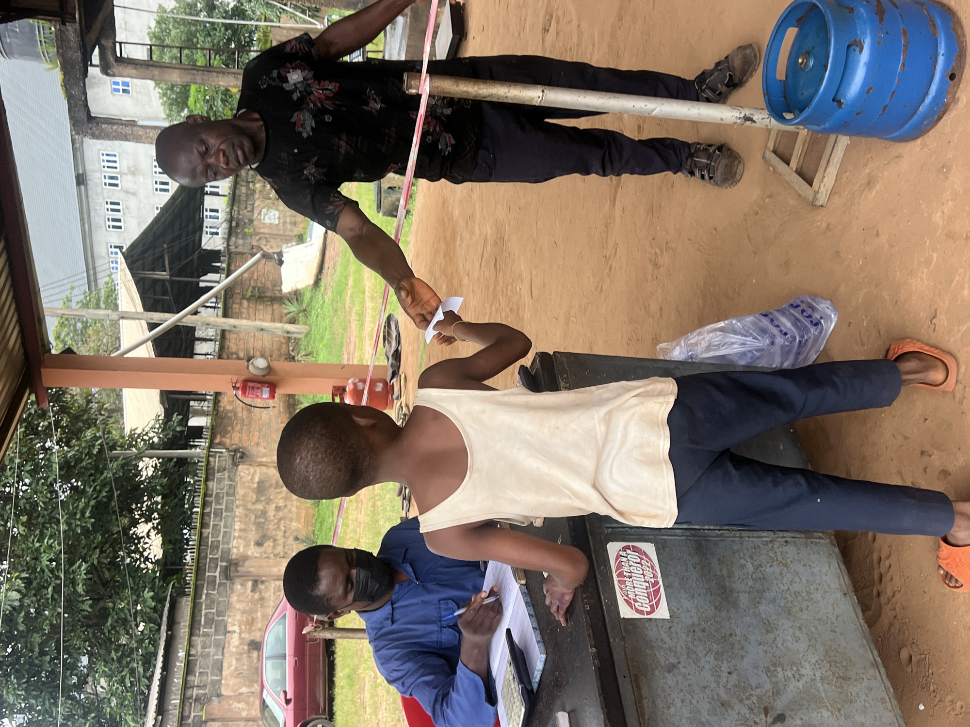 In Nkpor, Nigeria, a man wearing a blue uniform and a black face mask is seated behind a counter, writing on a ledger. He is holding a pen in his right hand. A boy wearing a white tank top and dark blue pants stands in front of him, offering a small white piece of paper. Another man, dressed in a black t-shirt with a floral pattern and dark pants, stands to the right, reaching out to the boy. A pink and red ribbon is stretched across the scene, separating the two men and the boy. In the background, a red car is parked, and buildings with multiple windows are visible. A blue gas cylinder sits on the ground to the right of the frame. The ground is covered in sand.