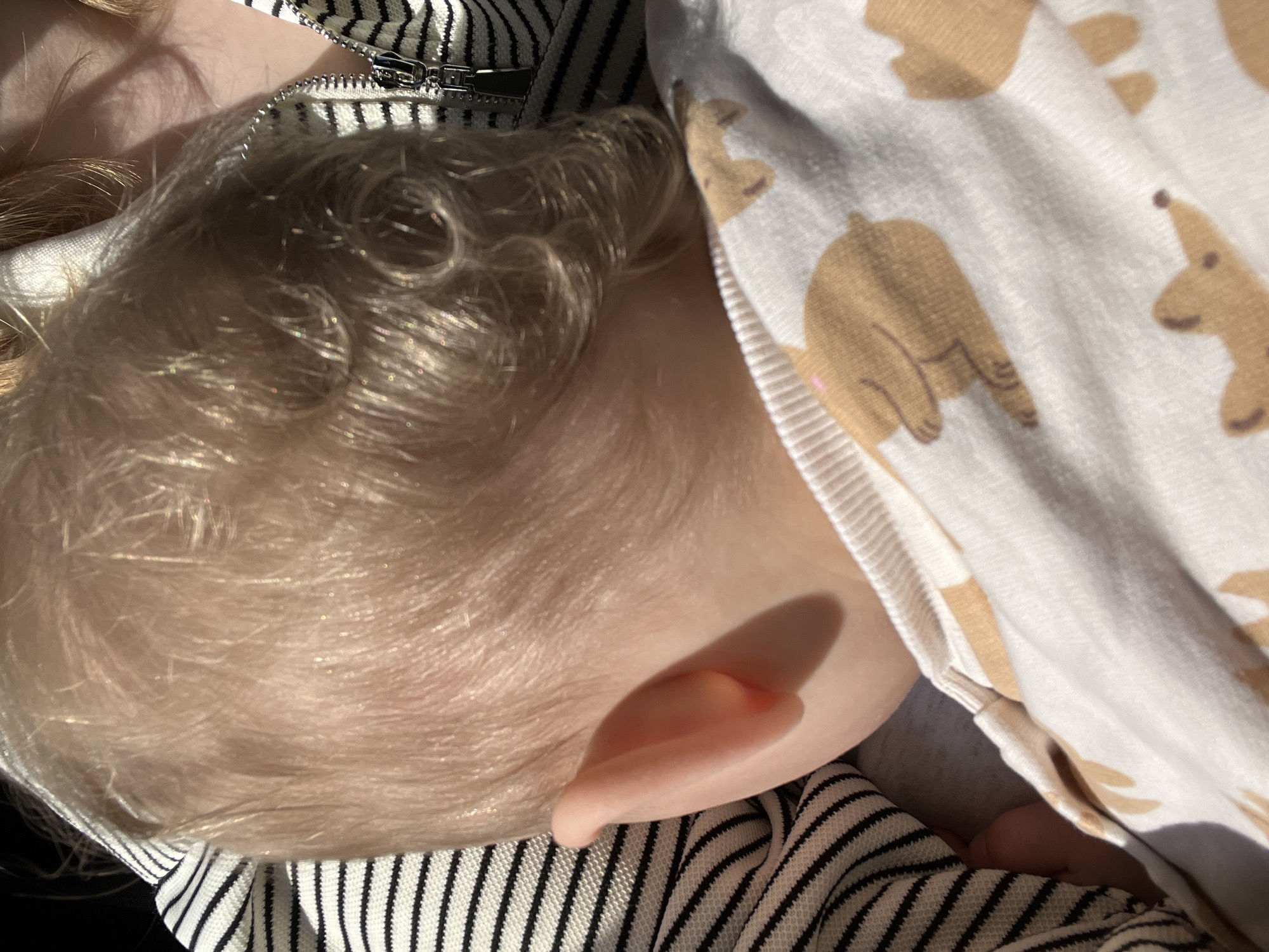 A close-up shot captures the side of a child's head, focusing on their blonde hair, ear, and partial face. The child is lying down, nestled among layers of clothing. A striped garment with a visible zipper detail is positioned on one side of the child's head, while a white fabric with brown bear patterns is draped on the other side. The scene is illuminated with natural sunlight, casting distinct shadows. The location of the city of Perm, Russia, is not detectable from the image alone.