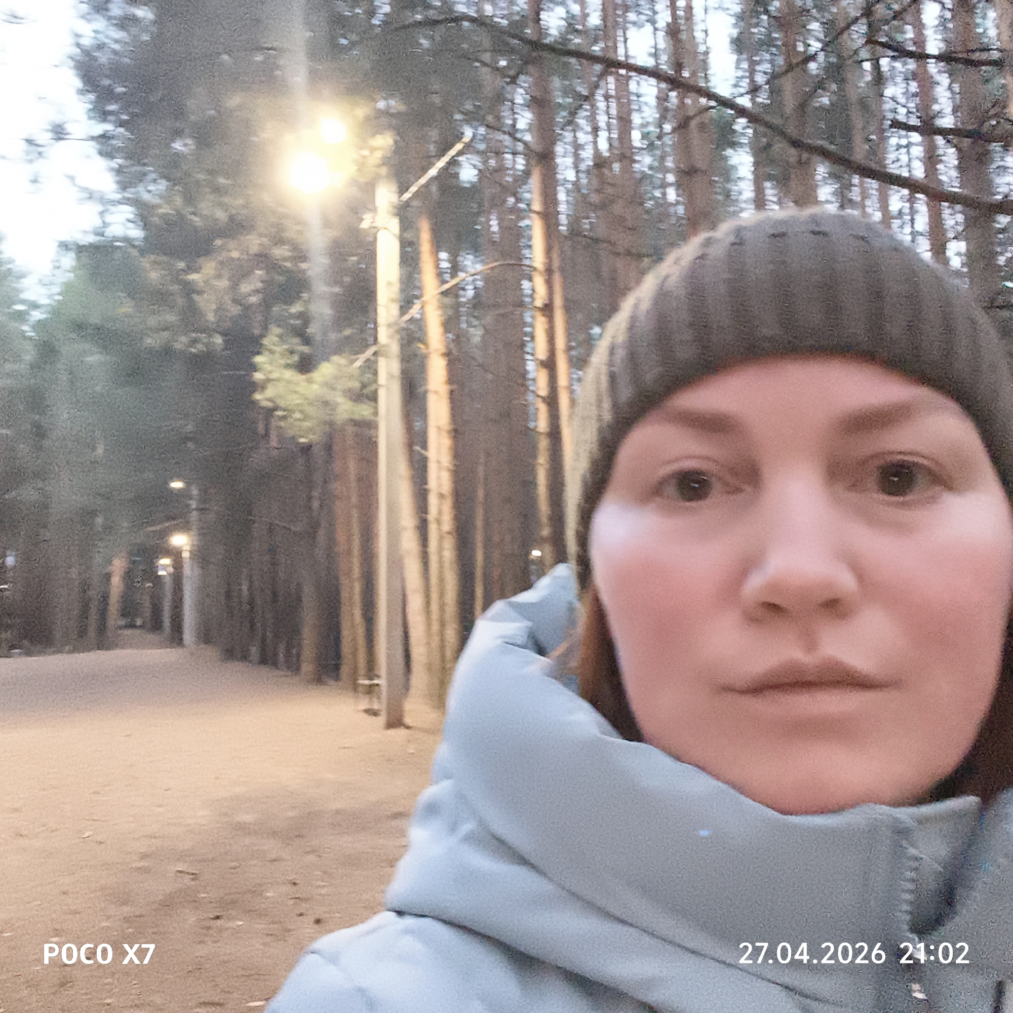 The image is a selfie taken by a woman outdoors on what appears to be a cool evening or night, likely in Chaikovsky, Russia. The woman, positioned in the foreground and slightly to the right, is wearing a dark knit beanie and a light blue or grey padded jacket. Her gaze is directed straight at the camera, and her expression is neutral.
The background shows a densely forested area, likely a park or a well-maintained walking path. Numerous tall, slender trees, resembling pines or other conifers, line both sides of a wide path that recedes into the distance. Several streetlights are visible along the path, casting warm, bright glows that illuminate the surroundings and create a sense of depth.
Notable details include the time and date "27.04.2026 21:02" displayed in the bottom right corner, confirming it is late evening. The device name "POCO X7" is visible in the bottom left corner. The woman's warm attire suggests cooler temperatures. The scene is tranquil, with the streetlights providing ample illumination despite the darkness, indicating a safe and accessible environment. No other people or distinct activities are visible in the frame.