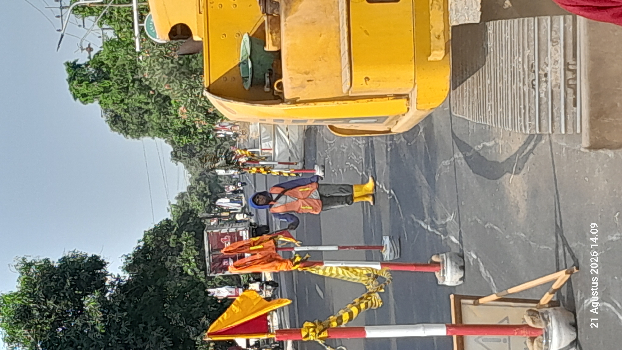 A person wearing a blue hooded jacket, an orange reflective vest, dark pants, and yellow boots stands on an asphalt road in Takeran, Indonesia. The individual is holding a red and yellow flag. To their right, a large yellow construction vehicle, likely an excavator, is partially visible, featuring a track and a green bucket. The road is marked with white and red striped poles, some adorned with yellow and black striped caution tape and orange flags. In the background, several trees line the road, along with a truck and motorcycles. The scene appears to be a construction site or roadwork area, with the ground showing signs of repair or modification. The timestamp "21 Agustus 2026 14.09" is visible in the lower left corner.