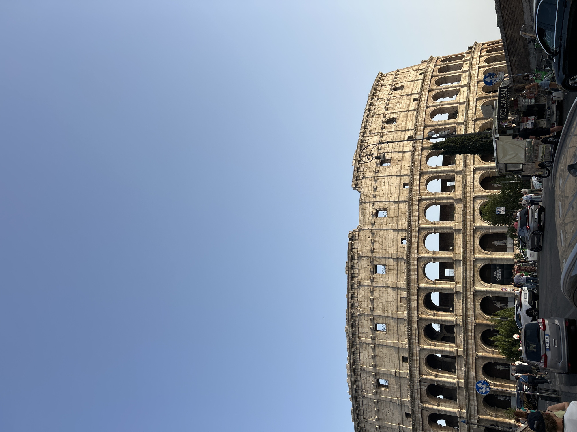 The image depicts the exterior of the Colosseum in Rome, Italy, under a clear blue sky. The ancient amphitheater dominates the upper two-thirds of the frame, showcasing its iconic arched facade and weathered stone. In the foreground, a street scene unfolds with several vehicles, including cars and an ice cream truck, and a number of pedestrians. The people are engaged in various activities, such as walking, standing, and interacting with the ice cream truck. The environment appears to be an urban setting, with a lamppost and some trees also visible. The lighting suggests it is daytime, with the sun casting shadows and highlighting the textures of the Colosseum.