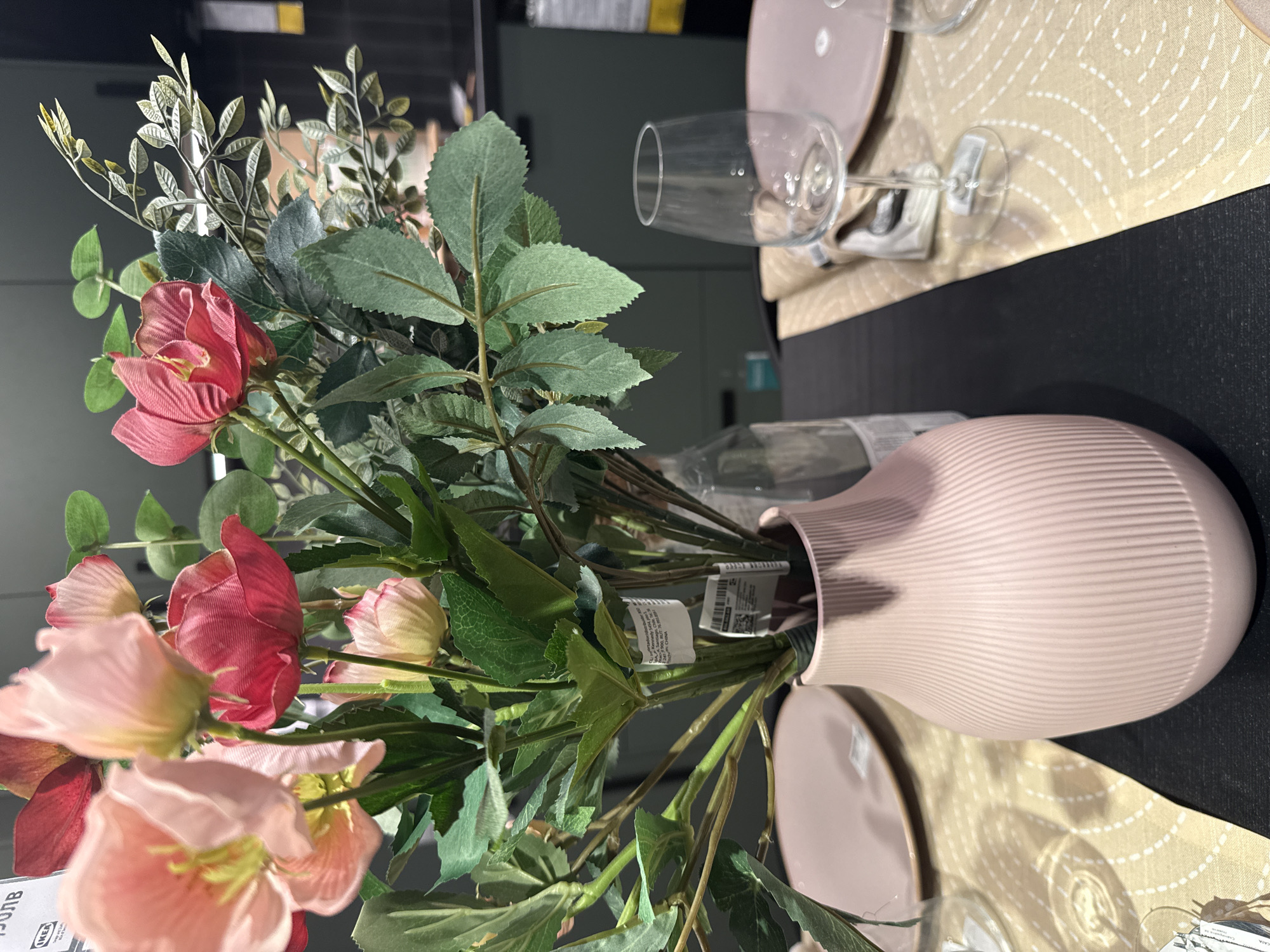 The image shows a bouquet of artificial flowers in a pink vase. The flowers are various shades of pink and red, interspersed with green leaves. The vase is cylindrical with a ribbed texture. The bouquet is placed on a table, with place settings nearby. The table is divided into a black section and a tan tablecloth section with a geometric pattern. There are pink plates and wine glasses on the tablecloth. The location is not evident from the image itself, and Sofia, Bulgaria, cannot be inferred from the visual information provided.