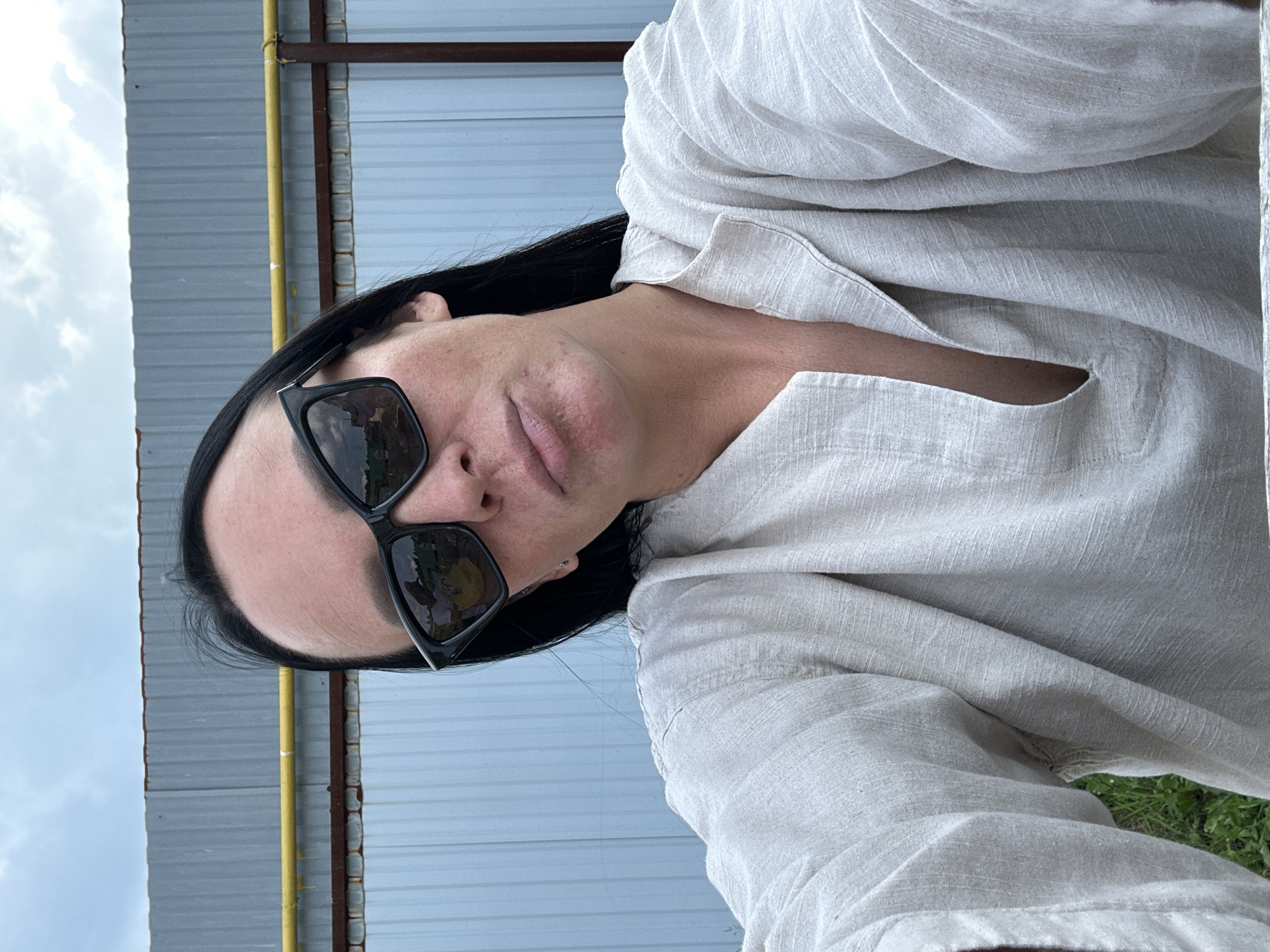 A woman with long, dark hair and sunglasses is taking a selfie outdoors. She is wearing a light-colored, loose-fitting shirt. Behind her is a corrugated metal fence with a yellow pipe running horizontally across it. The sky is cloudy. The background suggests an industrial or rural setting.