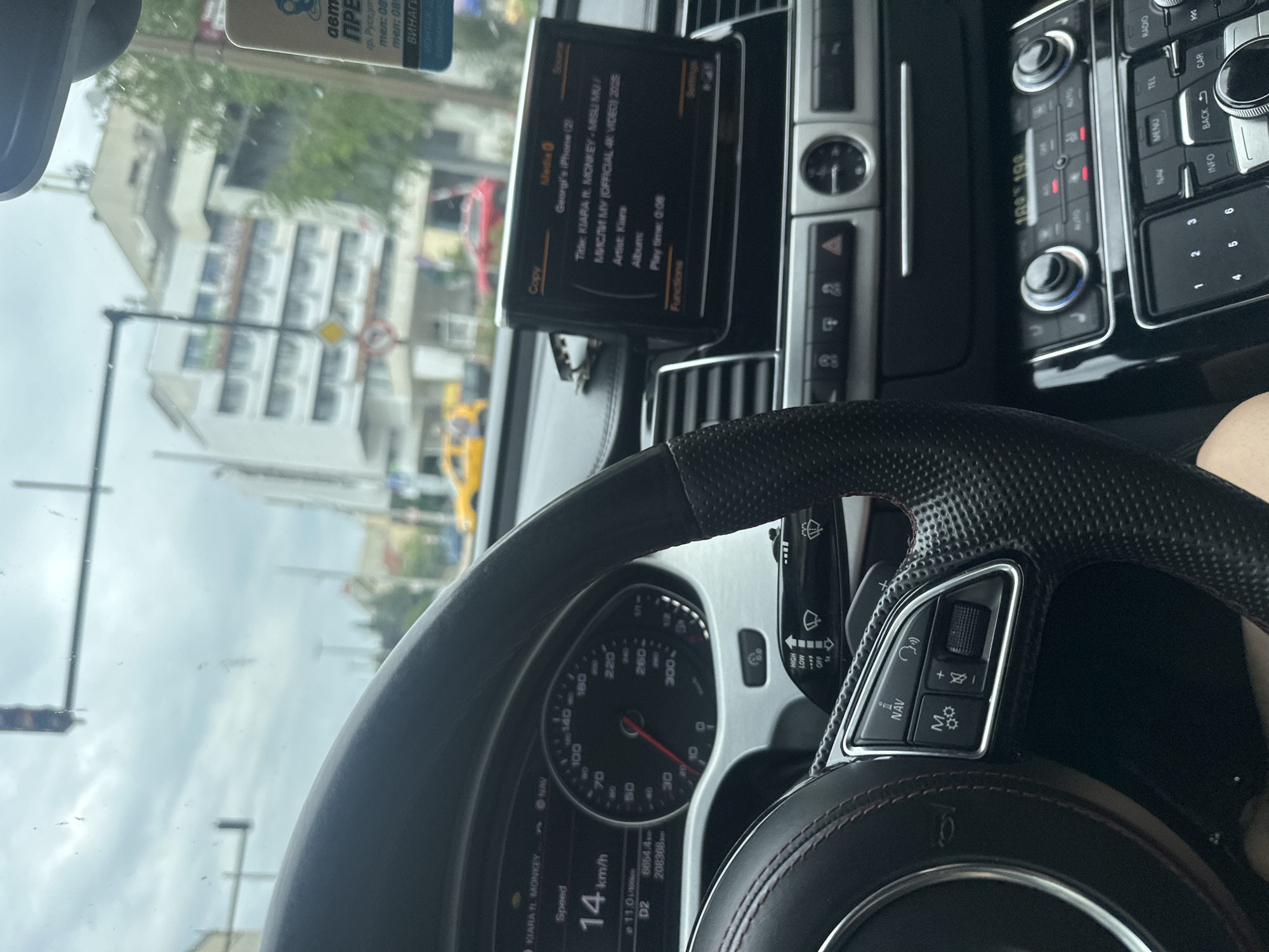 This is a media file showing the interior of a car, likely a luxury sedan, with a focus on the dashboard and steering wheel. The image is taken from the driver's perspective, looking out the windshield.
The primary subjects are the car's interior elements, including a digital display showing song information, a speedometer, and various controls for the car's infotainment system and climate control. The display shows "KIARA ft. MONKEY - MISU MU / МИСЛИ МУ (OFFICIAL 4K VIDEO) 2025" as the title of a song, with "Kiara" listed as the artist. The car's speed is indicated as 14 km/h.
Outside the car, through the windshield, we see a street scene. Buildings with multiple floors are visible, along with some trees and a bright yellow vehicle, possibly a taxi or public transport. Traffic signals and street signs are also present. The weather appears to be overcast or cloudy, judging by the diffused light and grey sky.
The setting is a street in Kazanlak, Bulgaria, as indicated by the text on a sticker visible on the windshield, which includes "гр. Русе, ул." and phone numbers. The presence of the song title and artist suggests that music is playing in the car, and the driver is likely on a journey. The overall mood conveyed by the image is one of a typical commute or drive through a city.
