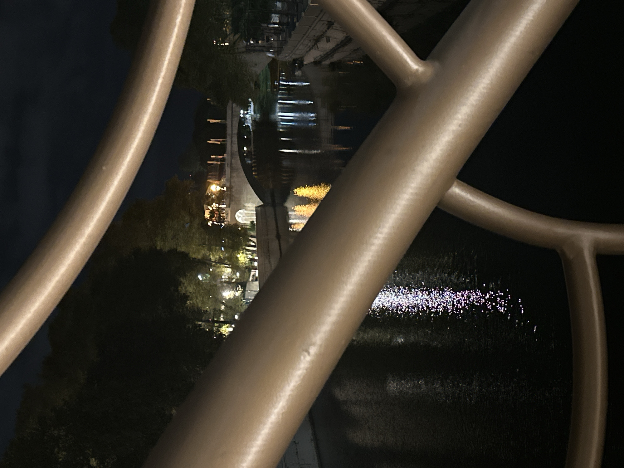 A nighttime view of the Sochi River embankment is captured through the railing of a bridge or walkway. The river's dark water reflects the lights from the surrounding cityscape and a distant bridge. Trees line the embankment on both sides, with illuminated buildings and streetlights visible amongst them. The foreground is dominated by the metallic railing, rendered in a bronze hue, which bisects the frame, creating a sense of depth. The overall atmosphere is serene and urban, highlighting the city's nighttime ambiance.