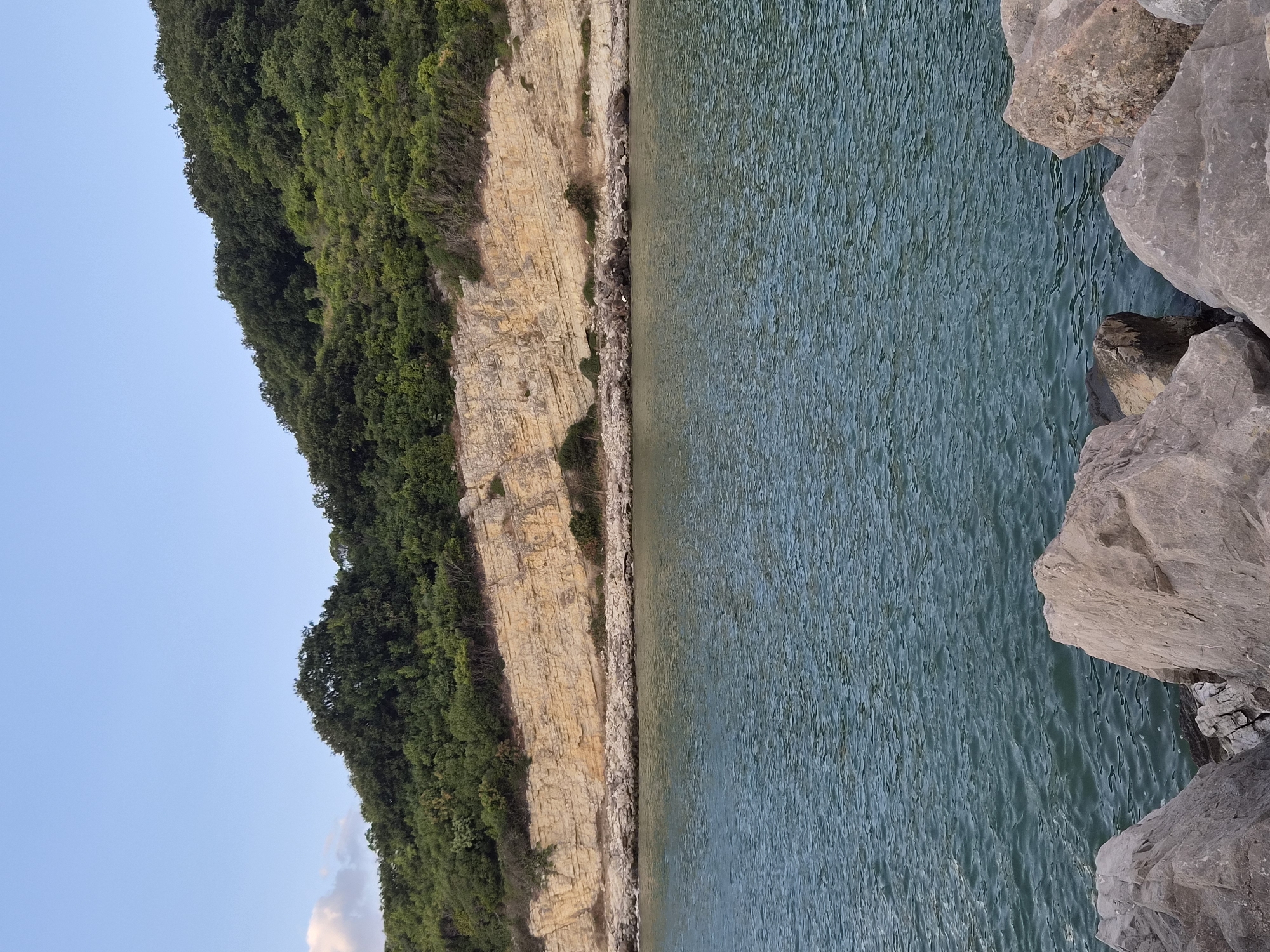 The image shows a coastal scene in Şile, Turkey. In the foreground, large, grey rocks are visible. The middle ground features a body of calm, turquoise water. Beyond the water, a rocky shoreline leads to a steep, layered cliff face of light brown rock. The cliff is partially covered with green vegetation. The background is dominated by a lush, green hillside covered in dense trees, sloping upwards towards a clear, pale blue sky with a few wispy clouds on the left. The overall impression is a natural, serene coastal landscape.
