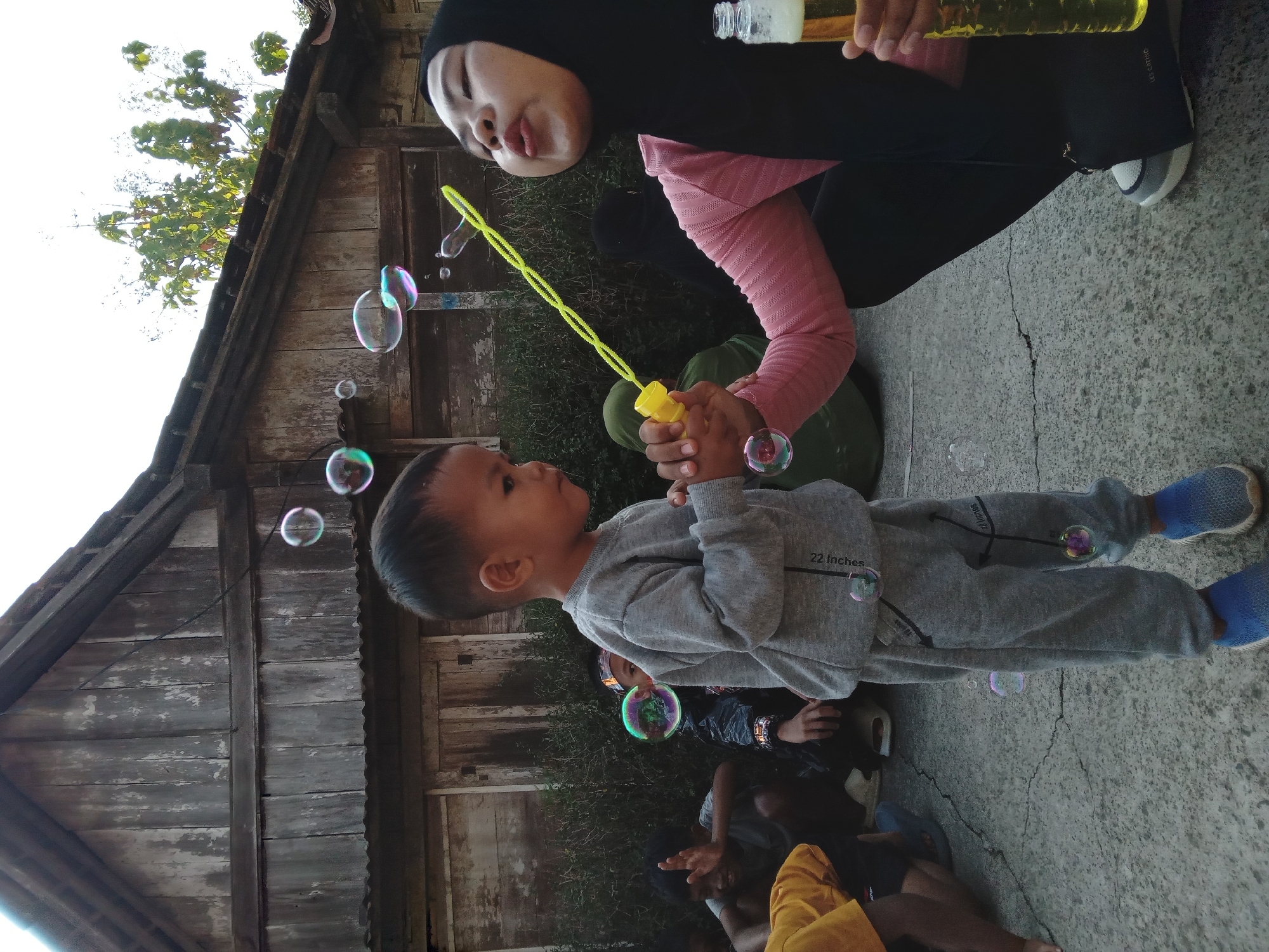 In the village of Kedunggudel, Indonesia, a young boy in a grey tracksuit stands facing right, looking at bubbles being blown by a woman wearing a black hijab and a pink long-sleeved shirt. She holds a yellow bubble wand connected to a container, and is actively blowing bubbles. She also holds a glass bottle containing a yellow liquid. Several bubbles are floating in the air around them. In the background, other people are visible sitting on the ground, and a rustic wooden building with a corrugated metal roof serves as the backdrop. The scene appears to be an outdoor gathering.