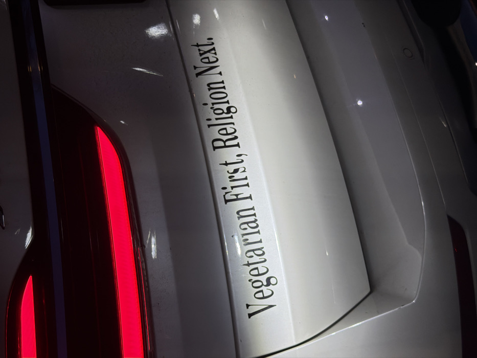 The image displays the rear section of a silver or light-gray vehicle, likely an SUV or crossover, observed in New York City, United States of America. A prominent black decal on the vehicle's tailgate reads, "Vegetarian First, Religion Next." The left taillight is illuminated with a horizontal red light strip. Above the taillight, a dark blue horizontal trim element is visible. The vehicle's paint surface exhibits several minor scratches and streaks of dirt. At least one circular parking sensor is visible on the lower right portion of the bumper. Multiple distinct reflections of artificial light sources are present on the glossy surface, suggesting an indoor or covered environment such as a parking garage. No individuals are depicted, nor are any direct actions or interactions.