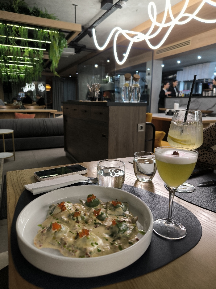 A close-up presents a white bowl filled with creamy ravioli topped with red caviar, accompanied by a green cocktail, two glasses of water, a smartphone, and a napkin on a round black mat. The setting suggests a restaurant with lush greenery and neon lighting, with glimpses of staff and other patrons in the background. The focus is on the meticulously plated food and drinks, inviting a sense of culinary enjoyment.