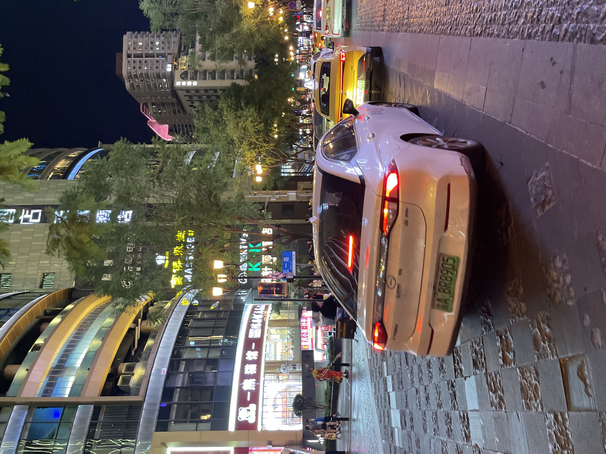 The image depicts a nighttime street scene in Yuzhong District, China, likely on a busy commercial thoroughfare. A white sedan is prominently featured in the foreground, with its taillights illuminated. Behind it, a yellow taxi with license plate A HA3077 is visible, followed by other vehicles, suggesting traffic. The street is paved with cobblestones and large rectangular tiles, some of which appear worn or damaged.
Buildings line the street, displaying bright neon signs and illuminated windows, indicating commercial establishments such as shops and possibly hotels. One sign with a bear logo and Chinese characters reads "一熊猫糖一" (Yi Xiong Mao Tang Yi), suggesting a candy or dessert shop. Another sign displays "KTV". Trees with illuminated streetlights dot the sidewalk. Pedestrians are seen on the sidewalk, including a person in an orange uniform carrying an object. The overall atmosphere is one of urban activity at night, with the characteristic blend of bright lights and darkness.