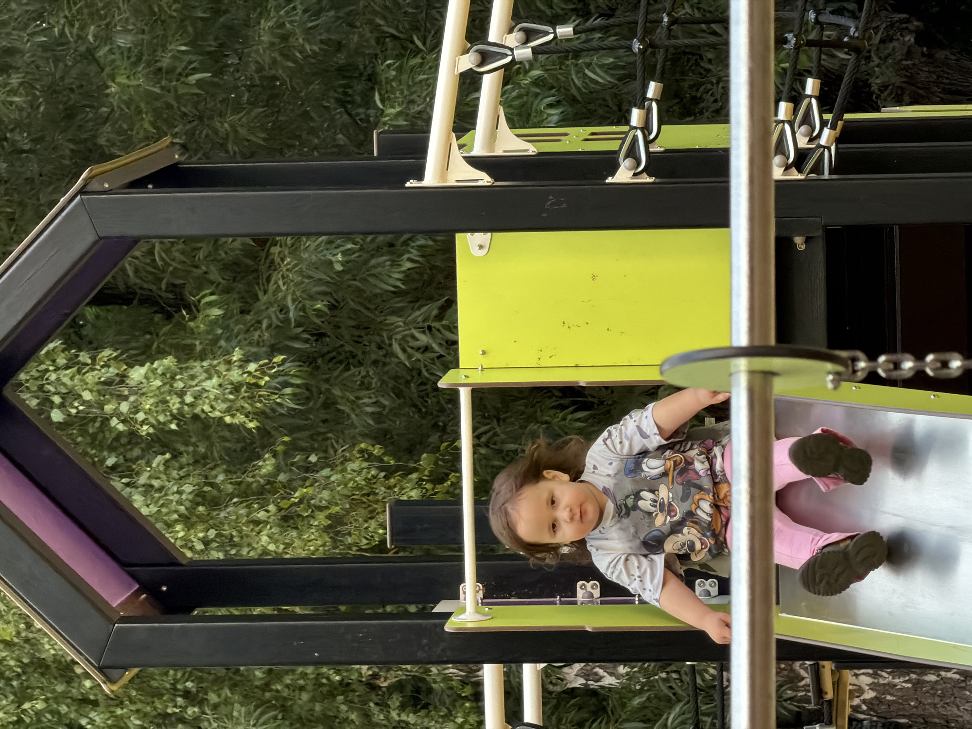 A young girl is sliding down a metal slide on a playground. She is wearing a gray t-shirt with images of cartoon characters and pink pants. Her feet are extended in front of her as she descends. The playground structure is made of black metal and bright green panels, with a purple roof. Green foliage and trees are visible in the background. The scene appears to be outdoors in a park.
