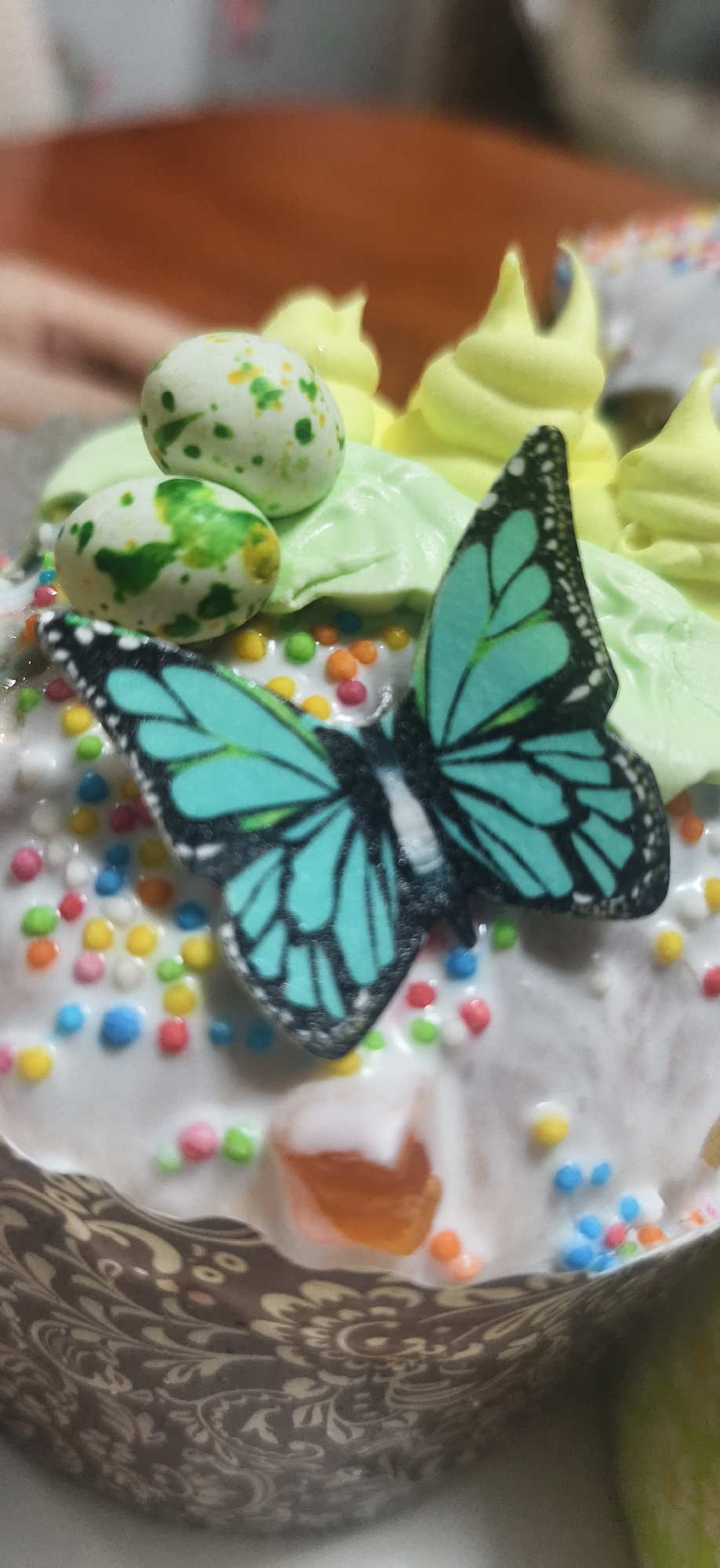 The image shows a decorated Easter cake. The cake is covered in white icing and sprinkled with colorful round sprinkles. A decorative butterfly sits atop the icing. There are also two speckled, egg-shaped candies and green and yellow frosting. The cake sits in a patterned paper cup. The background is blurred, showing a brown surface. There is no verifiable evidence to suggest the location is in the city of Фоки, Russia.