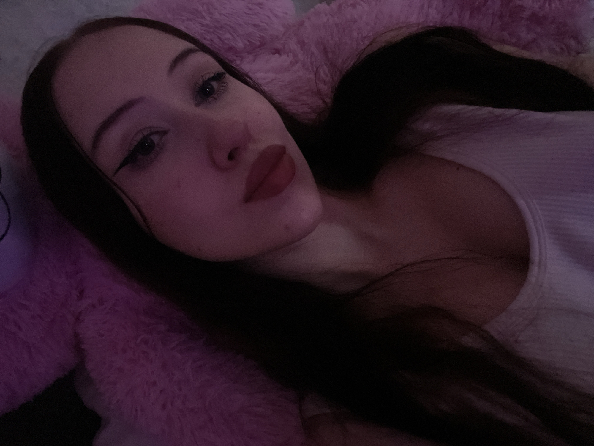 The media file is a close-up, indoor portrait of a young woman with long, dark hair. She is lying down, possibly in bed, with a soft, pink, plush surface behind and around her, which appears to be a blanket or a stuffed animal. She is looking towards the camera with a soft, relaxed expression, and her makeup includes eyeliner and lipstick. The lighting is dim, with purple and pink hues, suggesting it might be nighttime or a dimly lit room, possibly with colored lights. Her clothing is a light-colored, possibly white, ribbed tank top. There is no visible text in the image. The context of Votkinsk, Russia, provides a geographical location but no specific cues within the image itself.