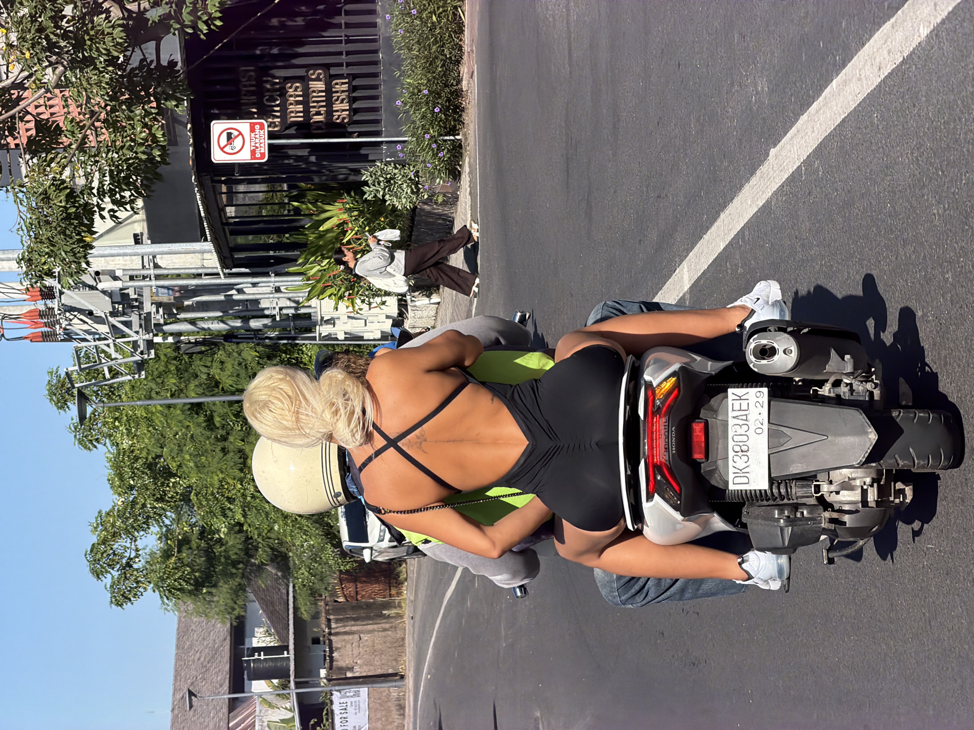 The scene captures a street in North Kuta, Indonesia, showcasing a man and a blonde woman riding a Honda motorcycle with the license plate "DK3803AEK 02.29". The woman is wearing a black, backless garment and white sneakers, while the man wears a helmet and jeans. The street is paved and marked with a white line. In the background, there's a building with a sign reading "TAPAS COCKTAILS SHISHA" and another sign with a red circle and diagonal line. A pedestrian walks alongside the building. The overall environment includes trees, a clear sky, and overhead power lines.