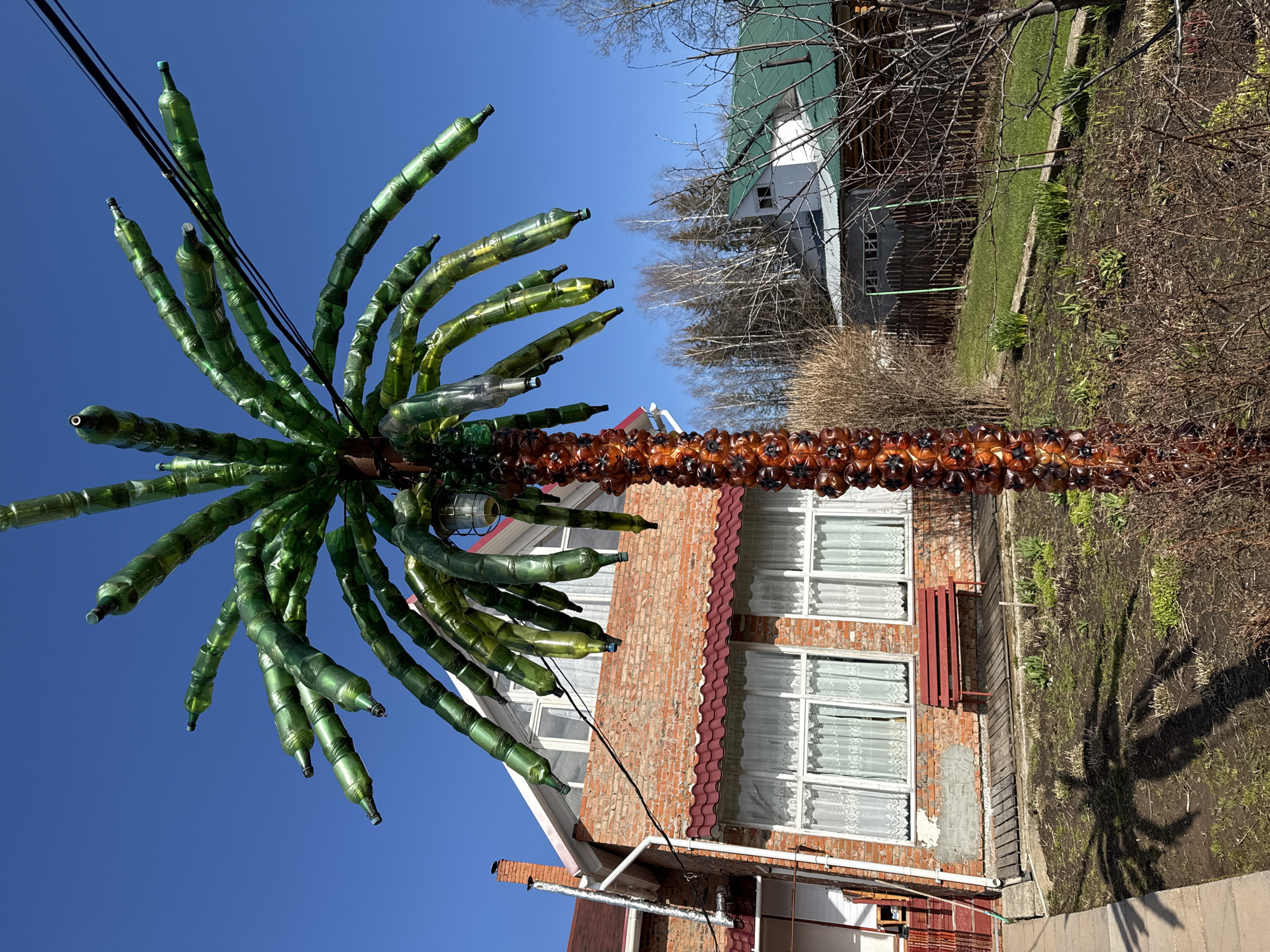The image shows a plastic bottle palm tree in front of a brick house. The palm tree's trunk is made of brown plastic bottles, while its leaves are made of green plastic bottles. The house has white-framed windows with lace curtains. A utility wire runs across the top left of the frame. The sky is blue. The surrounding environment includes a yard with bare patches and some greenery, and a neighboring house with a green roof.
While it is not possible to accurately identify the location of Adam, Russia, without additional information, the image provides a visual scene of a private residence with a handcrafted palm tree made of recycled materials.