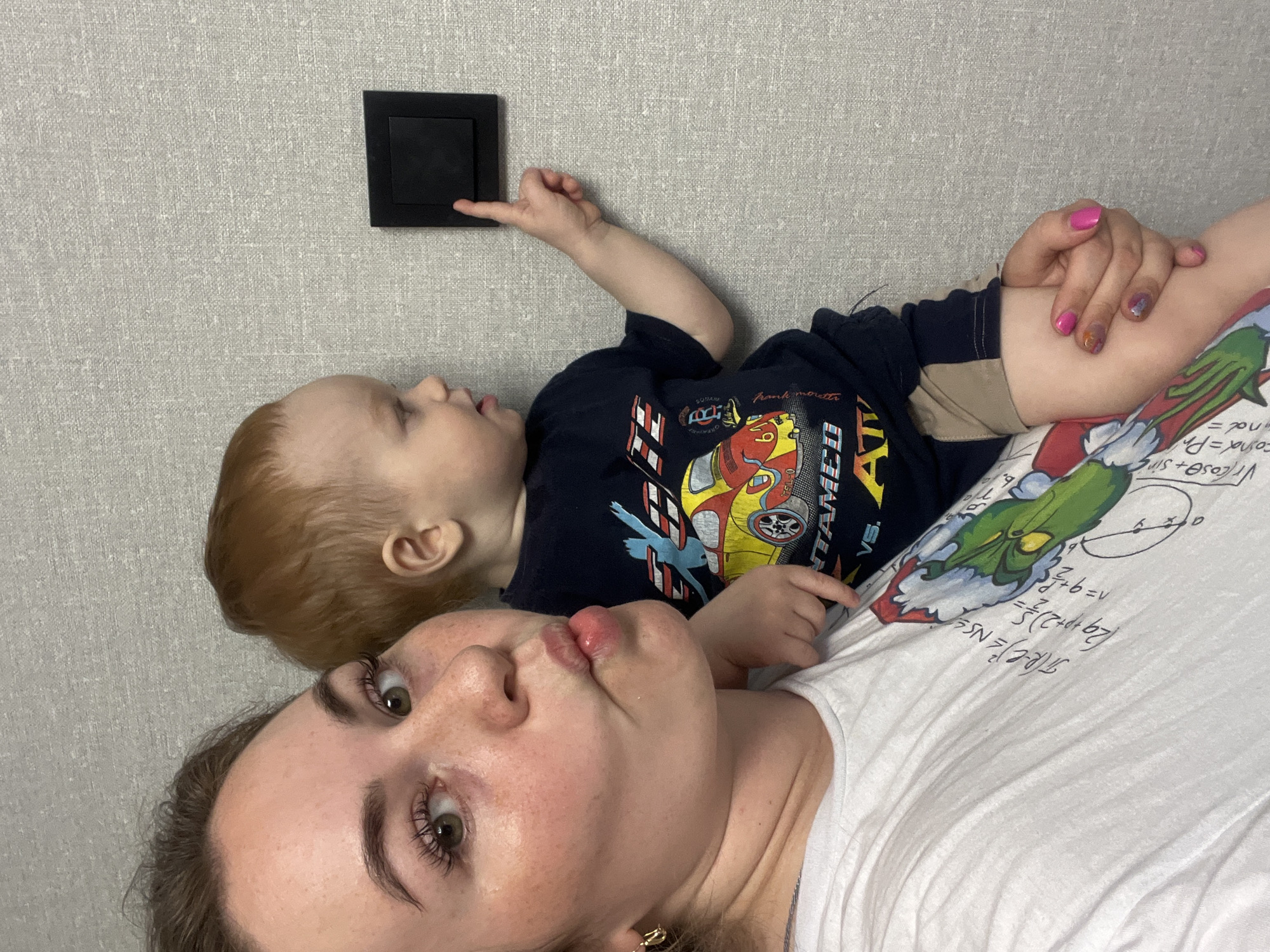 The image captures a playful moment between a woman and a young child in what appears to be an indoor setting in Chaikovsky, Russia. The subjects are positioned as if lying down, with the image rotated. The woman, presumably the mother, is in the foreground, facing the camera with a "fish face" expression, her lips puckered. She has long dark hair, freckles, and visible eyelashes. Her left hand shows nails painted in bright pink, with some iridescent or patterned nail polish on one finger. She wears a white t-shirt adorned with a green Grinch-like character and handwritten mathematical equations and symbols, including "R-CNS", "(29+p+2)=", "B", "37+6=v", "Vcosa+sin", "cosa = Pr", and "nd=".
Beside her, a young child with reddish-blonde hair lies with his head turned towards a light gray, textured wall. He is wearing a dark blue t-shirt with a colorful graphic featuring a racing car and various texts like "SONAM", "Rank monstr", "VS. ATV", "UNTAMED", "610", and "SL-0". His right index finger is actively touching a black light switch mounted on the wall, indicating curiosity or play. The overall scene conveys a relaxed and intimate interaction between the two individuals.