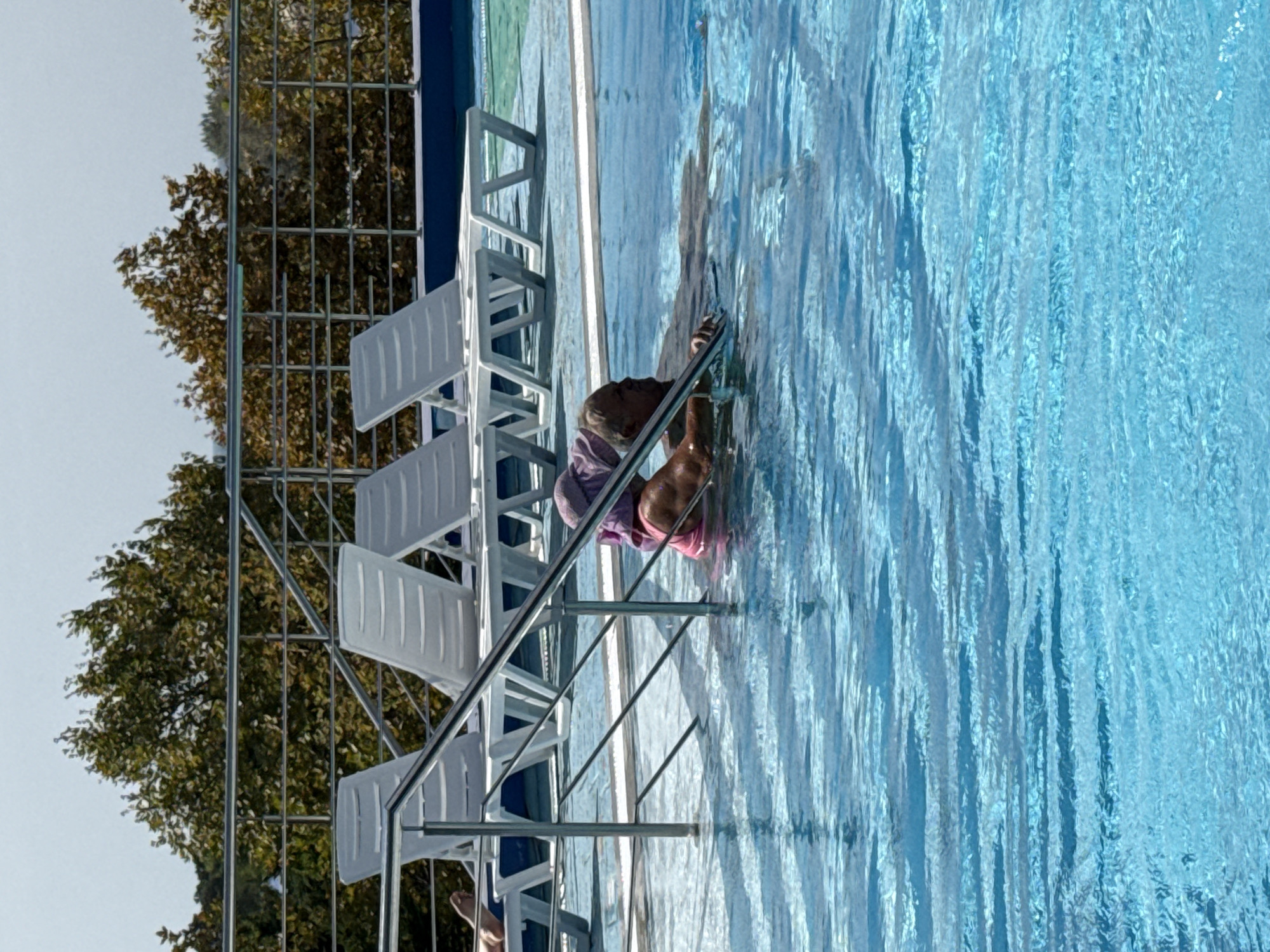 An elderly person with grey hair and a purple head covering is standing in a swimming pool, holding onto a metal railing. They are wearing a pink swimsuit and appear to be partially submerged in the bright blue water. Behind them, several white lounge chairs are arranged on the pool deck, which is bordered by a metal railing. Trees with green and brown foliage are visible in the background, and a clear sky is overhead. The location is the city of Sevastopol, Ukraine.