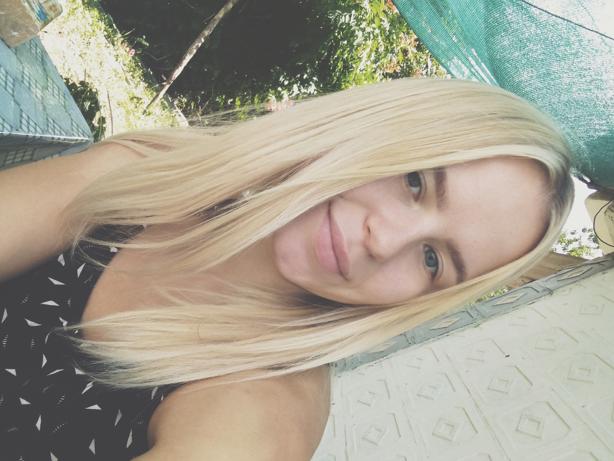 In Radisne, Ukraine, a young woman with blonde hair poses for a selfie outdoors. She is wearing a black top with a white geometric pattern. Behind her, a green mesh shade is visible, along with lush green foliage and a tree branch. To her right, a wall with a diamond-shaped pattern provides a textured background. The scene is illuminated by natural light.