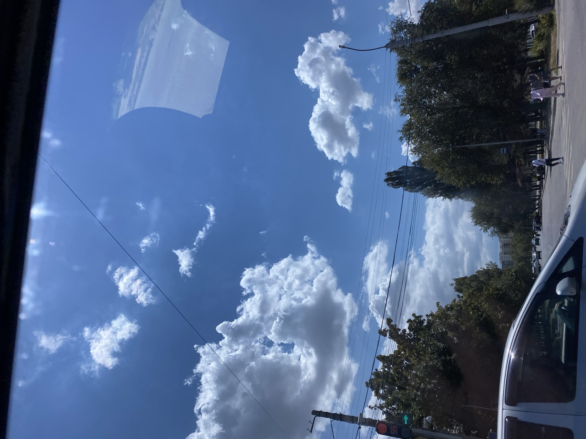 The image captures a street scene in the city of Bishkek, Kyrgyzstan, under a bright blue sky dotted with fluffy white clouds. In the foreground, the side of a silver vehicle is visible, indicating the perspective is from inside a car. A traffic light on the left shows a green arrow, with the number '55' displayed below the red light. Tall, leafy trees line the street on both sides, with a few taller, more slender trees interspersed. In the distance, buildings can be seen, suggesting an urban environment. A few pedestrians are crossing the street at a crosswalk, and other vehicles are present on the road, indicating moderate traffic. The overall atmosphere is that of a sunny day in a bustling city.