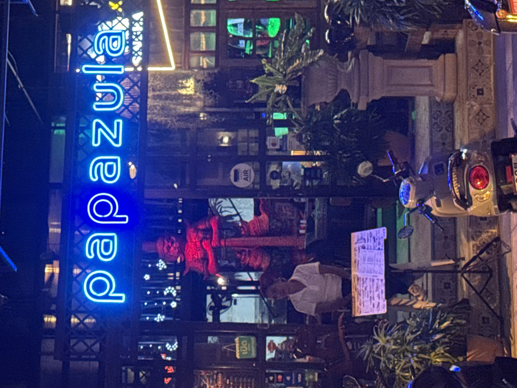 The image shows a nighttime scene outside a business identified by a bright blue neon sign that reads "PAPAZULA." The sign is mounted above an entranceway which is illuminated by warm yellow and white lights. To the left of the entrance, a large, red, statue-like figure, possibly a guardian or deity, stands holding a long staff. In front of the statue and to the left of the entrance, a person wearing a white polo shirt is seated behind a stand displaying a menu or information. In the foreground, a scooter is parked, and to the right, another scooter is partially visible. The environment appears to be a street or walkway with potted plants and architectural features suggesting an establishment in Phuket City Municipality, Thailand.
