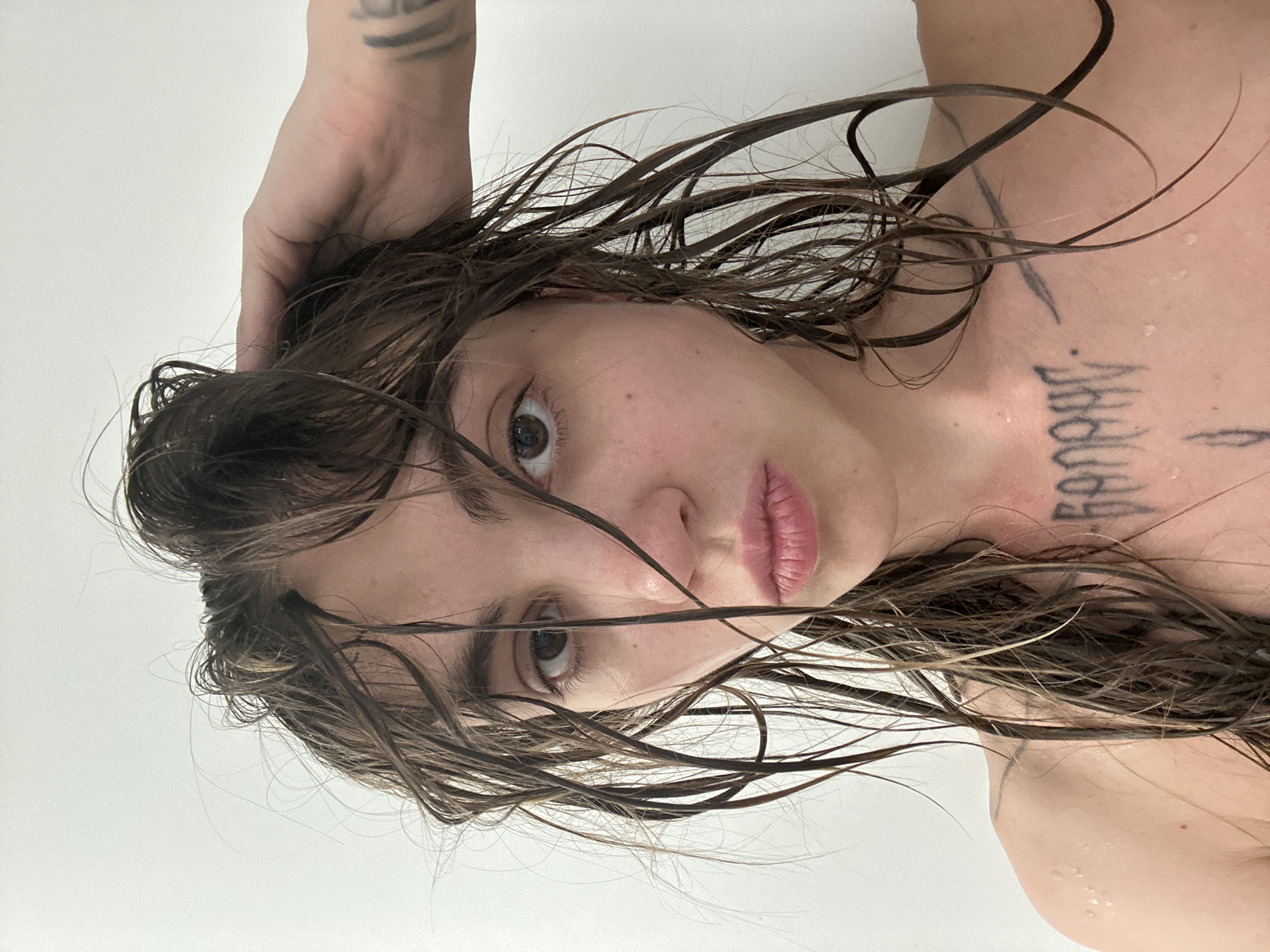A young Caucasian woman with dark brown hair, wet and tousled, is shown from the chest up in a selfie taken indoors against a plain white wall. She is looking directly at the camera with a neutral expression. Her left hand is raised and touching her hair. Water droplets are visible on her skin, particularly on her shoulders and upper chest. She has tattoos on her left forearm, upper left chest, and sternum. The lighting suggests a bright, indoor environment, possibly a bathroom. No specific location in Moscow, Russia, can be identified from the image.