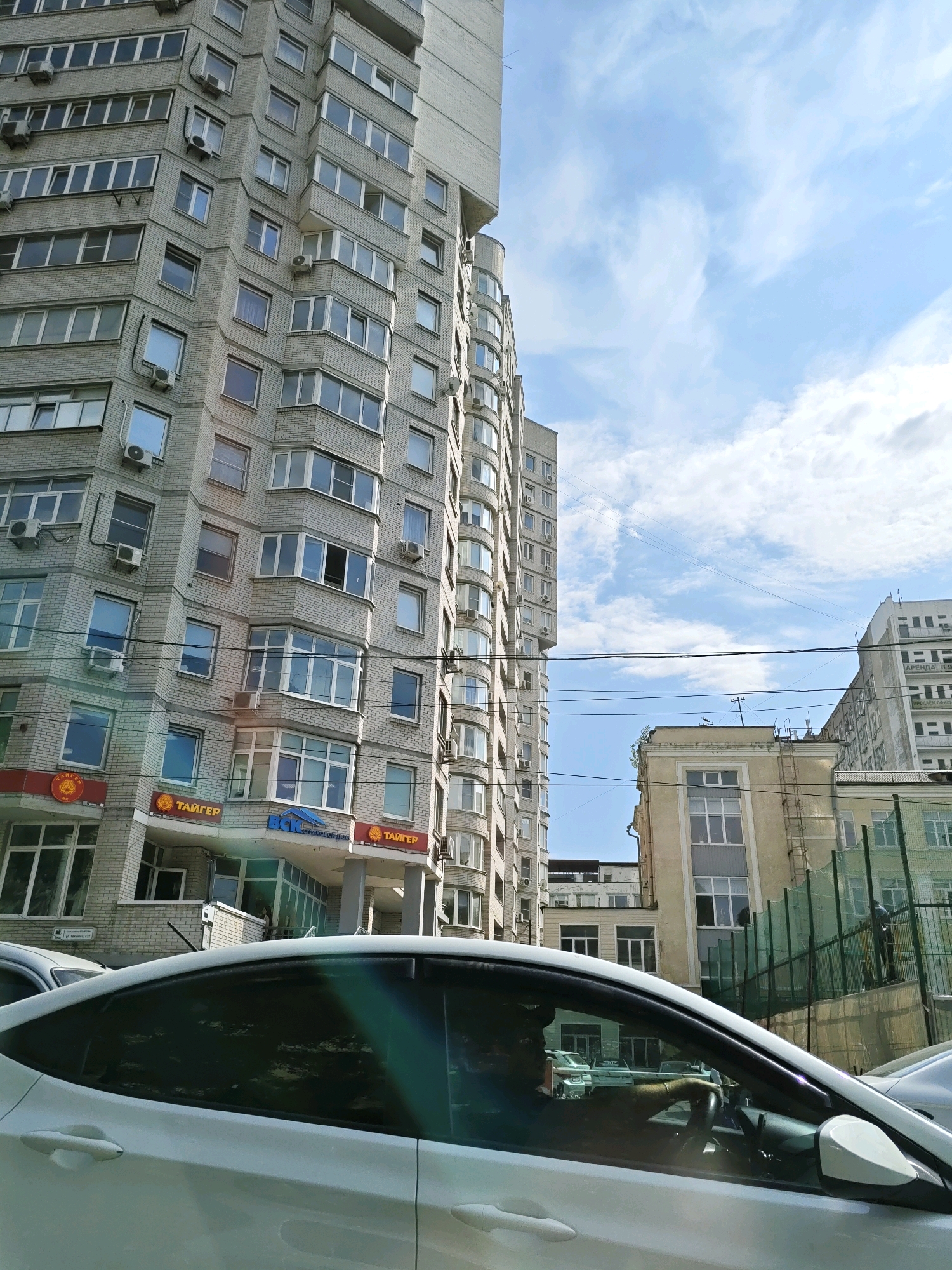 A side view shows the interior of a white car parked on a street in Rostov-on-Don, Russia, with a person visible driving. The street is lined with modern, multi-story buildings constructed with light-colored brick and featuring numerous windows and air conditioning units. Some of the buildings have businesses on the ground floor, indicated by signage such as "ТАЙГЕР" and "ВСК". Overhead, power lines crisscross the sky, which is partly cloudy with a clear blue sky visible. In the background, other buildings and a fenced sports court are present. The overall scene depicts a typical urban environment on a sunny day.