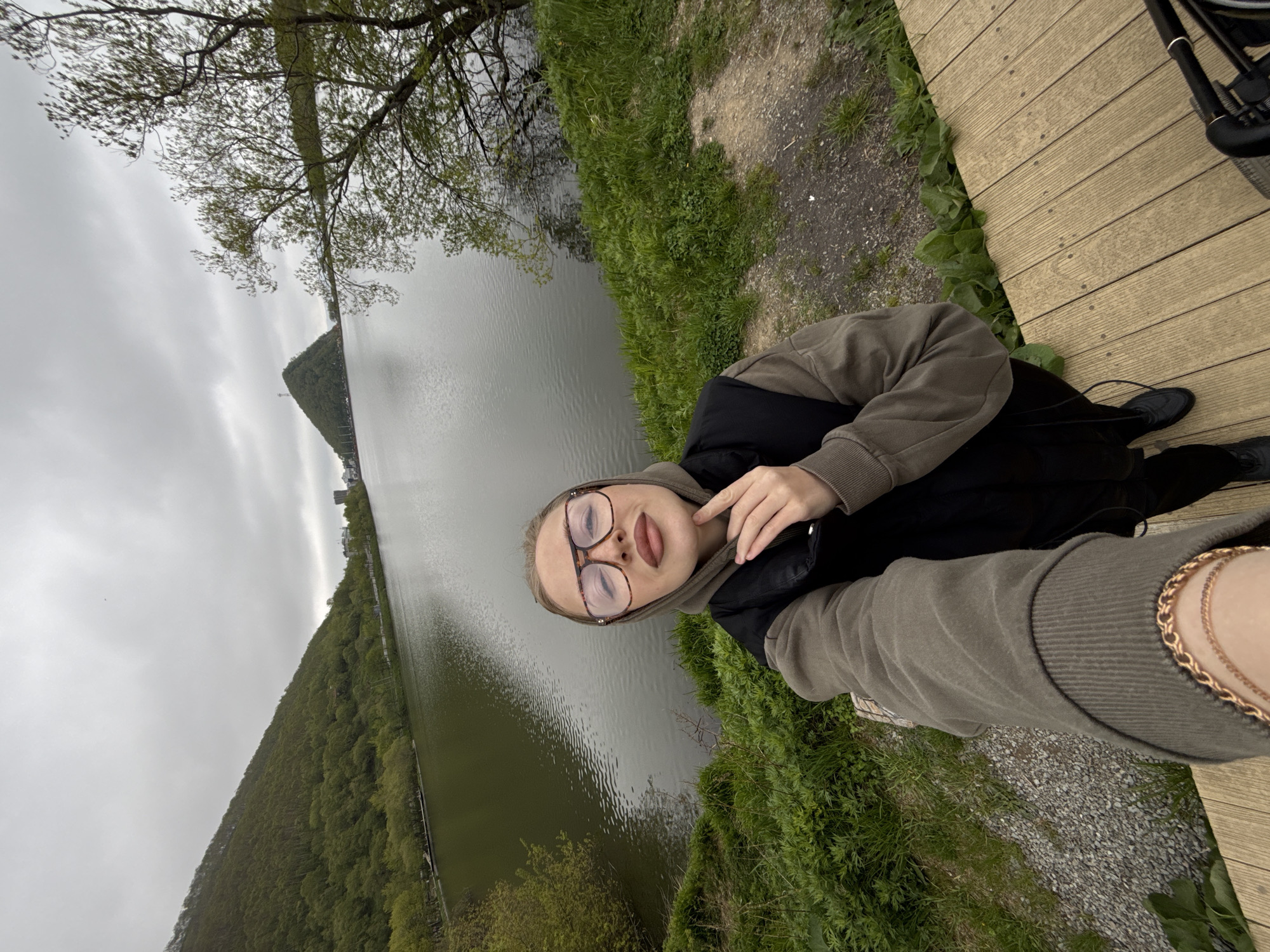A selfie is taken by a young woman with blonde hair, wearing large-framed glasses, a green hooded sweatshirt, and a black vest. She is standing on a wooden deck next to a body of water. In the background, there are green, tree-covered hills under a cloudy sky, and a mountain with a pointed structure on top. The scene appears to be a scenic outdoor location, possibly a park or recreational area, within or near the city of Petropavlovsk-Kamchatsky, Russia, given the distinctive conical mountain in the background, which resembles Mishennaya Mountain. The woman is touching her chin with her right hand, looking towards the camera with her lips slightly parted.