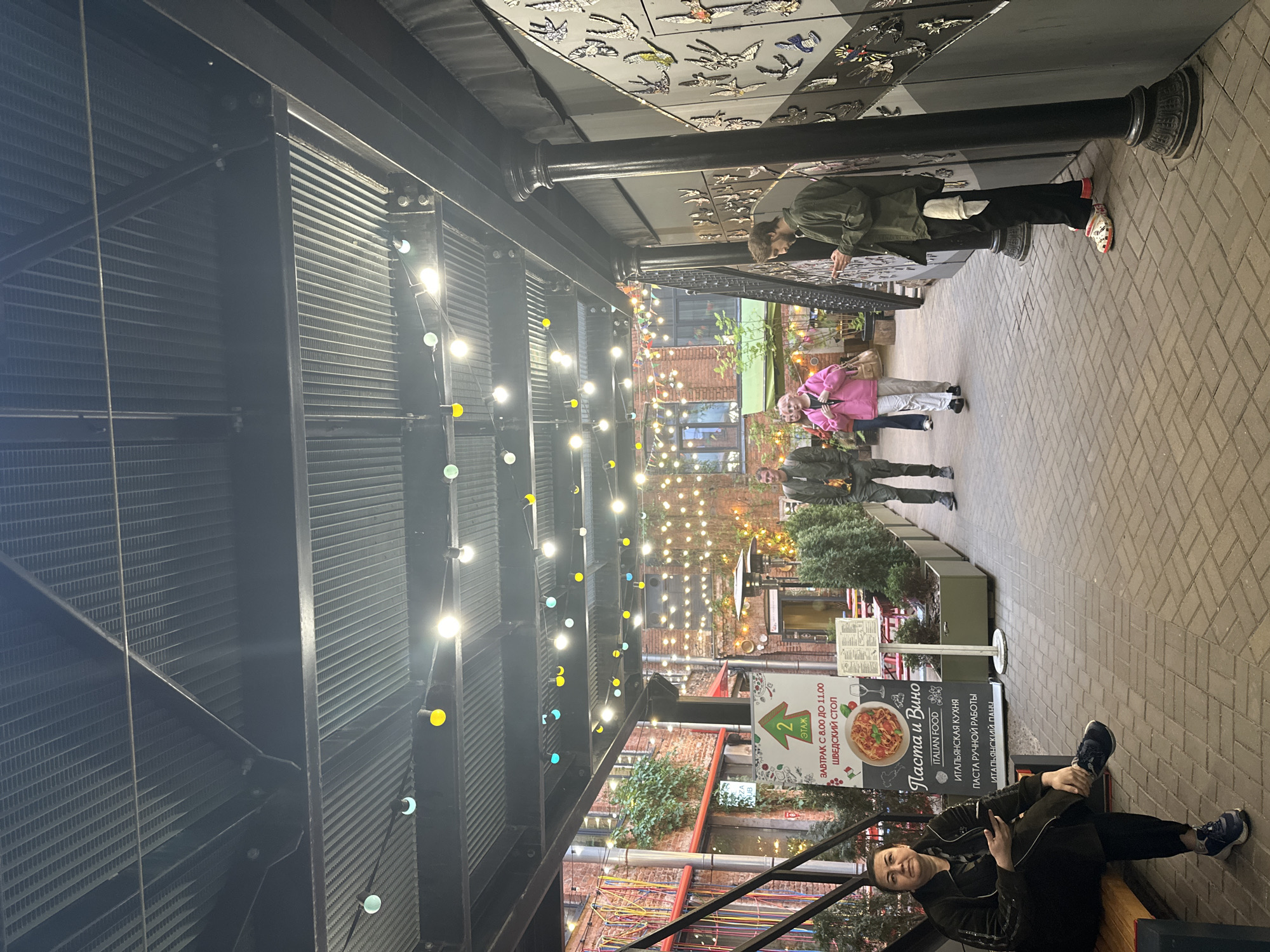 The image captures a scene within a covered walkway or courtyard in Saint Petersburg, Russia. The space is decorated with string lights in various colors hanging overhead, illuminating the area. The architectural elements include a metal frame structure supporting the roof, corrugated metal panels, and brick buildings visible in the background.
Several people are present in the scene. In the foreground, a woman sits on a bench, facing the camera. Further back, two people are walking along the brick-paved path. Near the entrance to the courtyard, another person is looking at something mounted on the wall.
Signage is visible, advertising "Pasta and Wine" in both Russian and English, indicating an Italian food establishment. The signs also mention breakfast being served until 11:00 am.