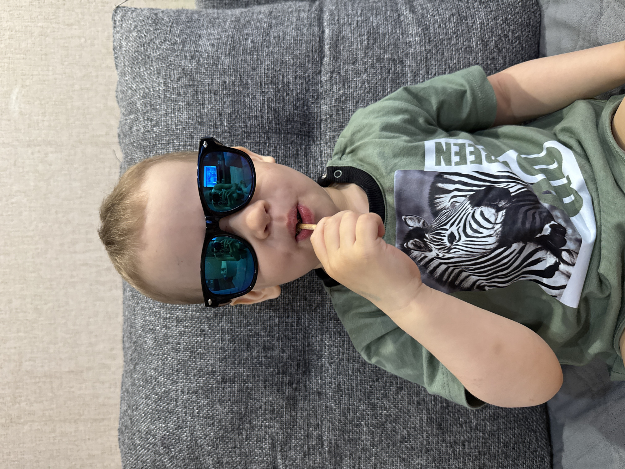 A young child is depicted lying down on a grey, textured surface, likely a sofa or cushion, in an indoor setting. The child has light-colored hair and is wearing black sunglasses with blue mirrored lenses. Reflections in the lenses show a room's interior, possibly including a screen or window with a blue light source, and what appears to be part of a person's legs or a reflection of the child's own lower body.
The child has a small, light-colored, stick-like object, possibly a snack or a toy, held between their lips. They are dressed in a green t-shirt featuring a prominent white graphic of a zebra's head on the front. Above the zebra, the word "GREEN" is visible in white capital letters. Due to the child's relaxed, sideway orientation in the image, the text appears upside down from the viewer's perspective. The child's left arm is bent, with their hand near their mouth. To the left of the child and the sofa, a lighter, subtly textured wall or floor surface is visible. The overall scene suggests a casual and relaxed moment, possibly playtime or a break. The location context provided is Rusanovo, Russia, though no specific visual cues within the image confirm this.