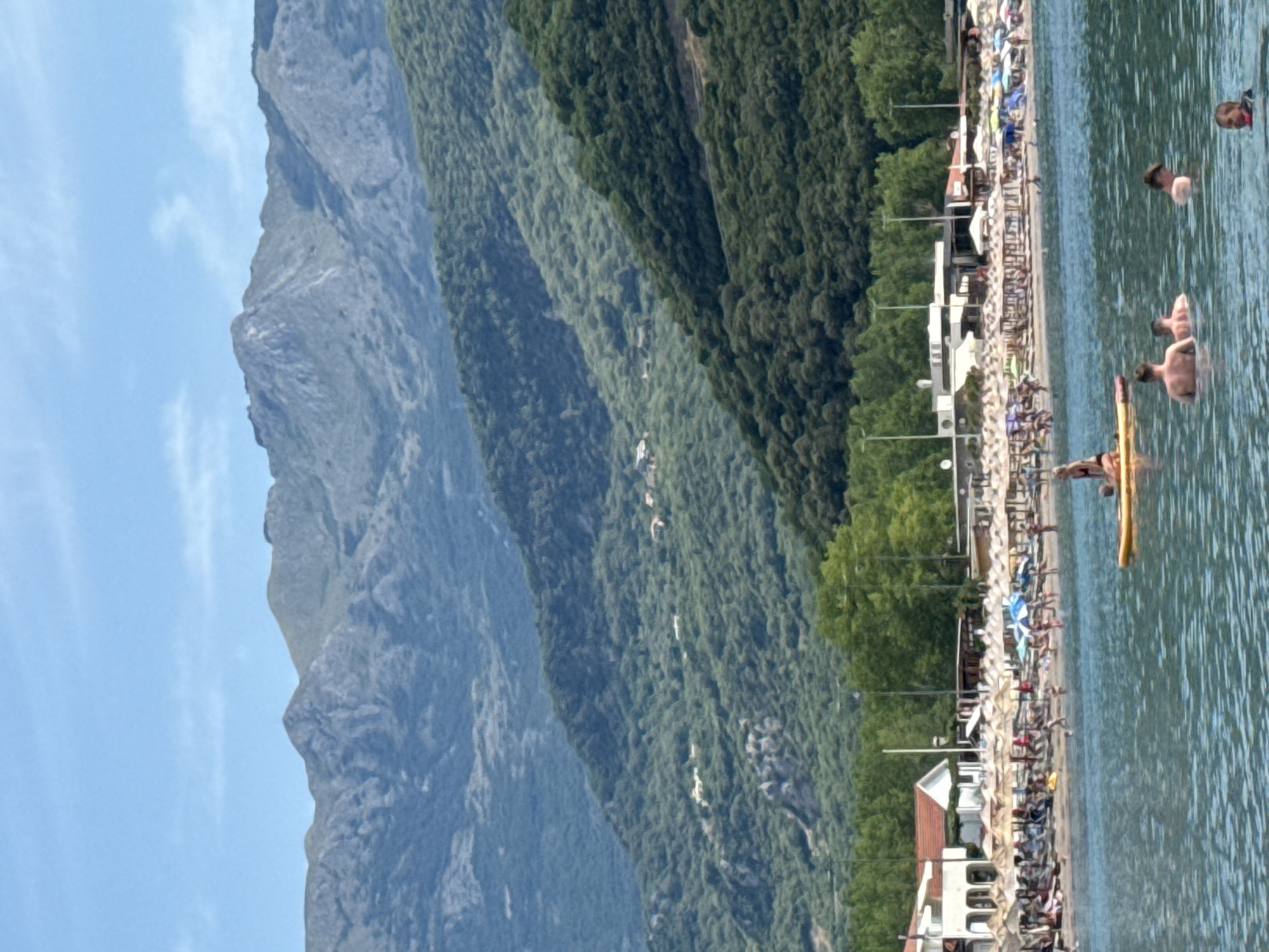 The image depicts a coastal scene with a mountainous backdrop, likely in Prijevor, Montenegro. In the foreground, the clear blue water is populated by several individuals. Two men are partially submerged, facing away from the camera, while another man is also in the water. A person is seated on a yellow paddleboard, and a woman is partially visible near them.
The middle ground reveals a bustling beach with numerous umbrellas and lounge chairs set up on the sand. Several people are visible on the beach, some appearing to be sunbathing or walking. Buildings, possibly restaurants or beach facilities, are situated behind the beach, characterized by white facades and red roofs. Trees line the area behind the buildings.
The background is dominated by lush, green mountains that rise steeply. The lower slopes are densely covered in trees, with some scattered houses or structures visible among them. The upper parts of the mountains are rocky and bare, with a rugged texture. The sky is a clear, bright blue with faint white clouds. The overall impression is a vibrant summer day at a popular seaside resort.